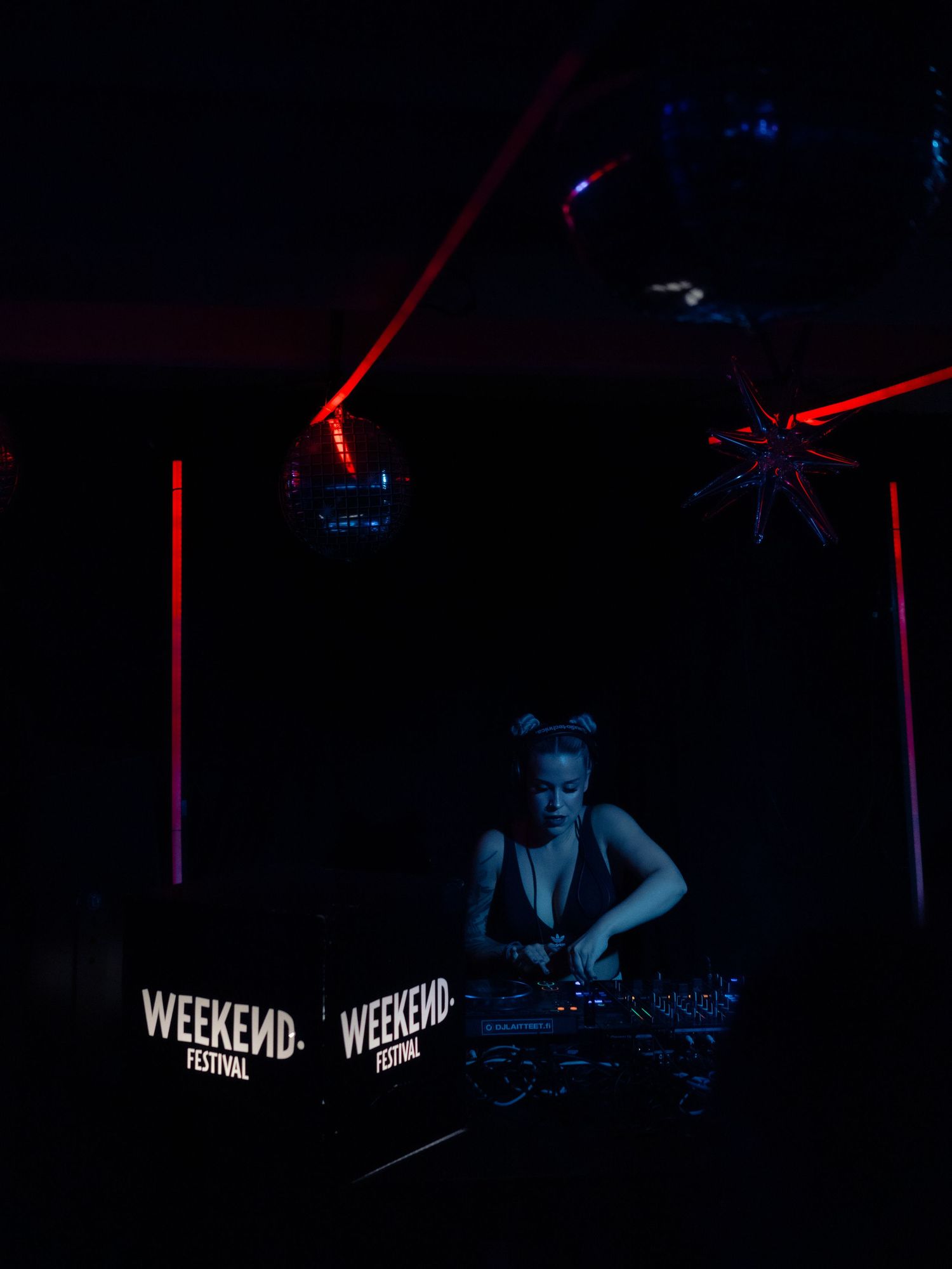 A DJ performs at a mixing console under red lighting with Weekend branding visible in the dark nightclub atmosphere.