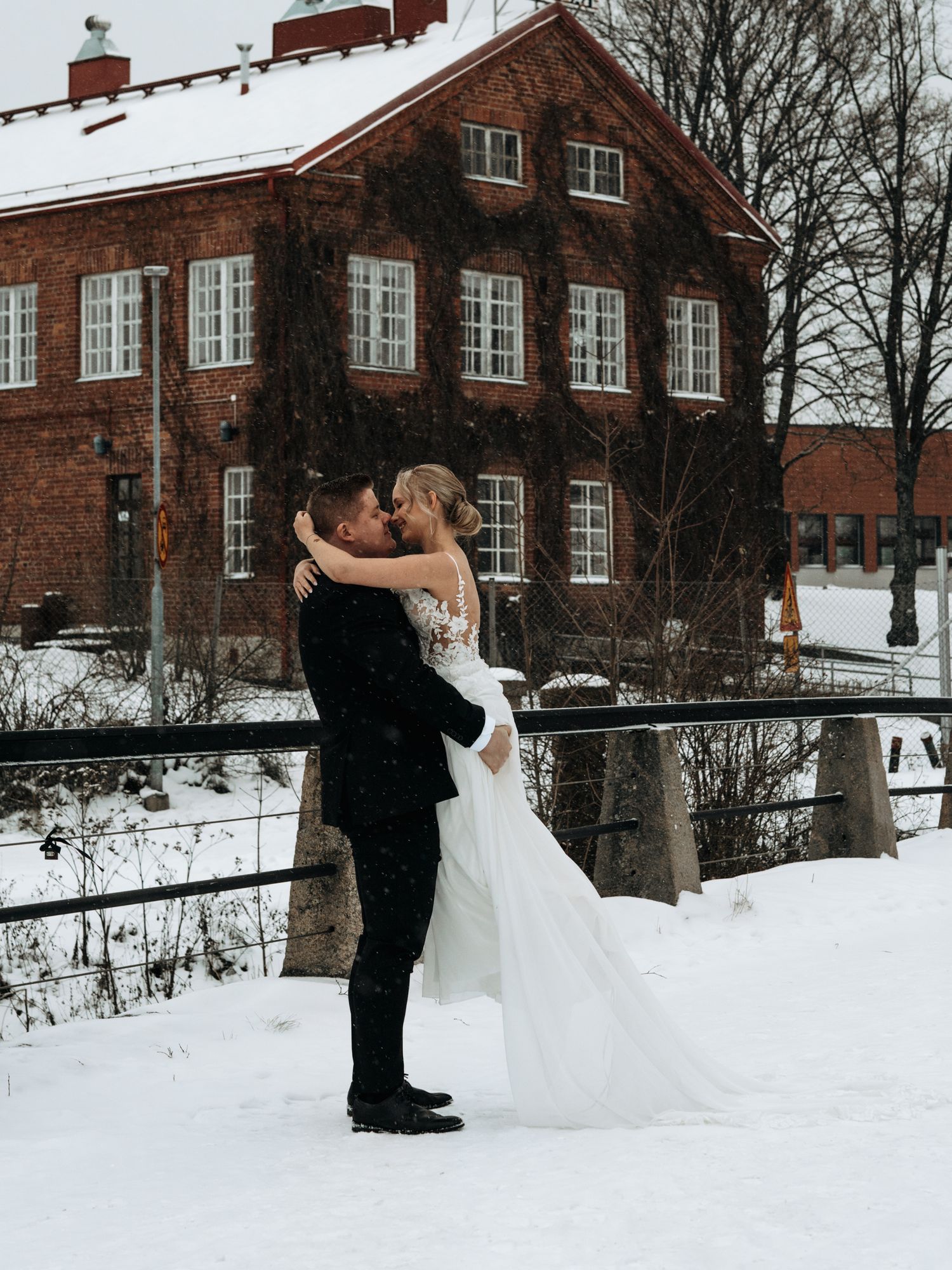 A romantic winter wedding scene with a couple embracing outside a snow-covered brick building in Nordic style architecture.