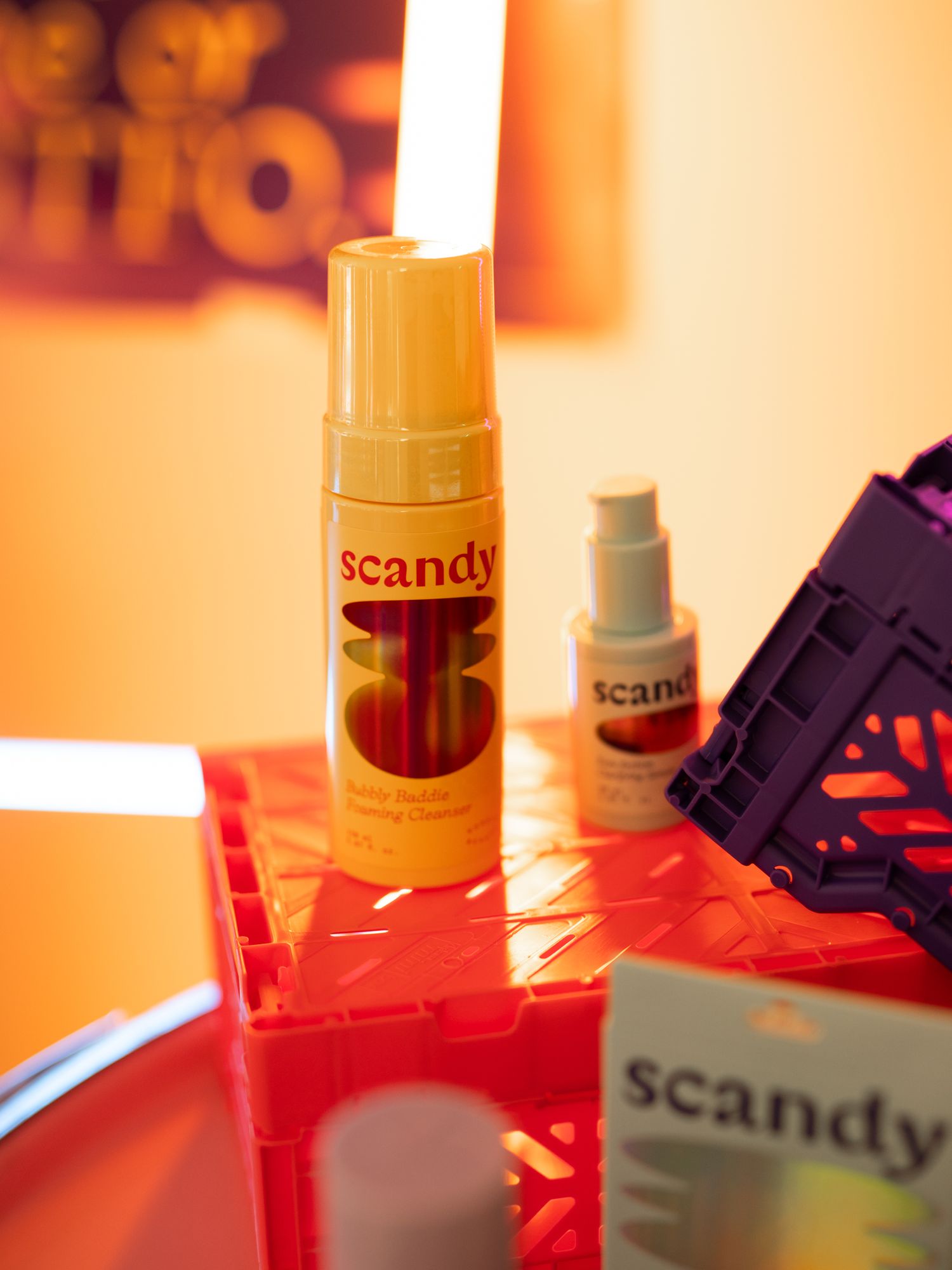 Scandy brand beauty products displayed on a red reflective surface with orange ambient lighting.