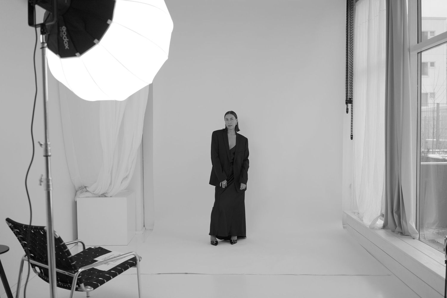A black and white studio photo showing a figure in a black suit standing in front of a white backdrop with lighting equipment.