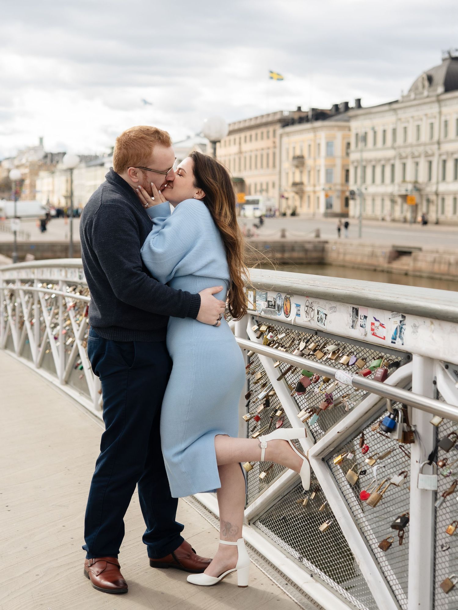 Couple sharing a romantic kiss on a bridge with love locks, city buildings visible in the background.