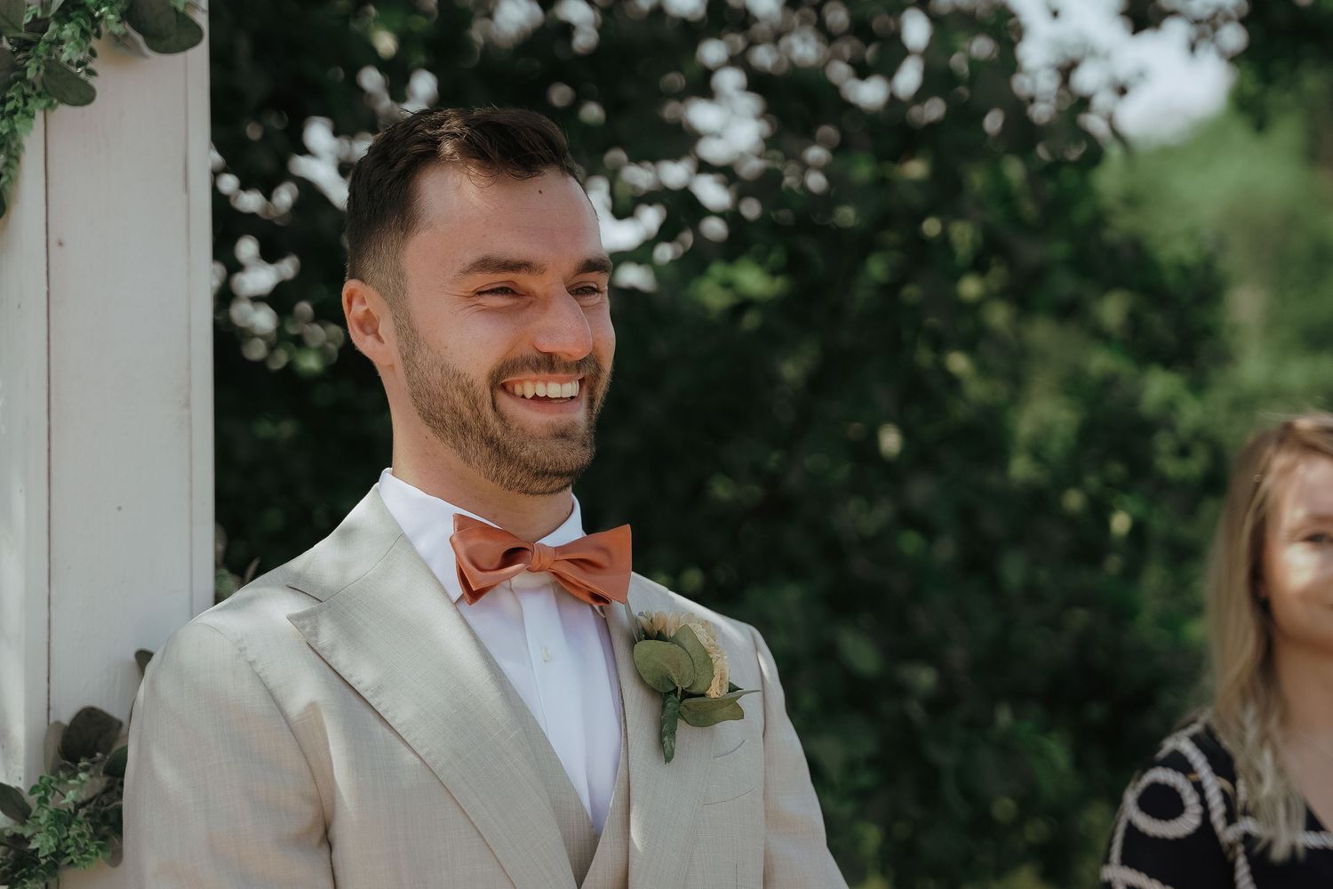 Groom smiling in light gray suit with coral bow tie and boutonniere in an outdoor garden setting.