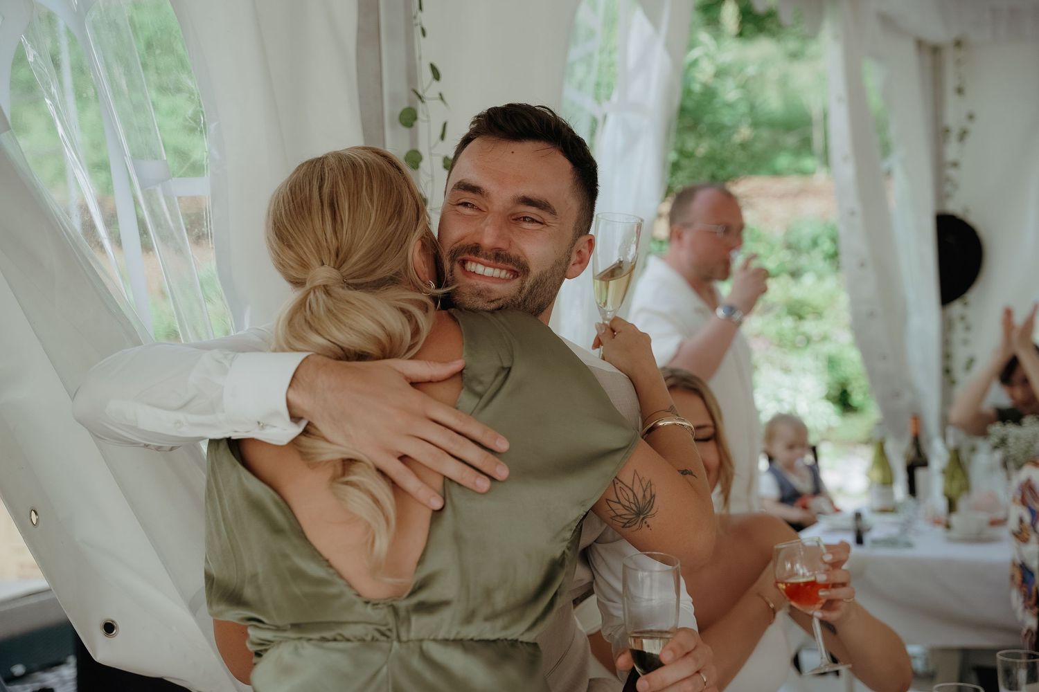 A joyful embrace at a wedding reception under a white tent, with guests visible in the background.