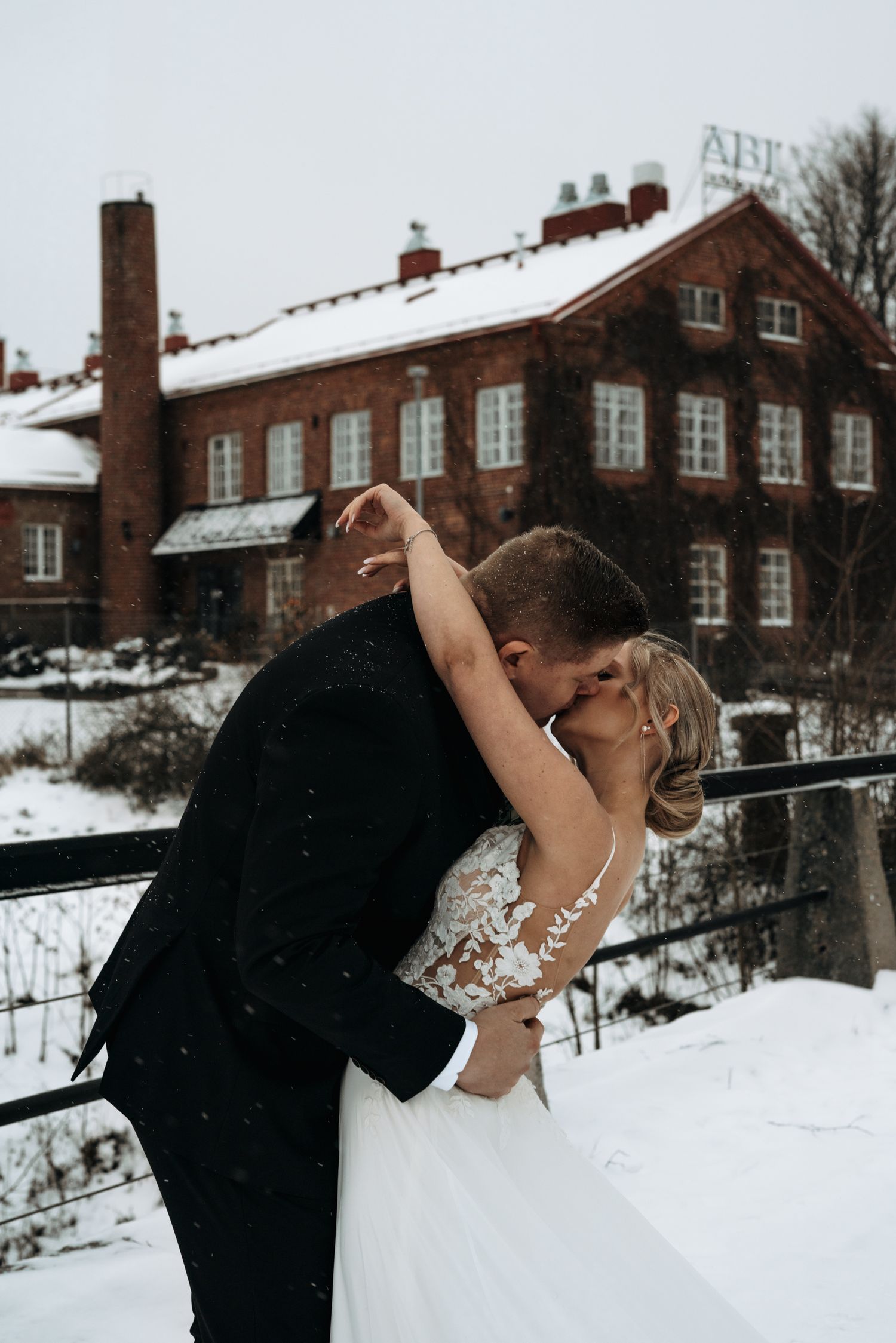 A romantic winter wedding photo showing a couple embracing in front of a snow-covered brick building.
