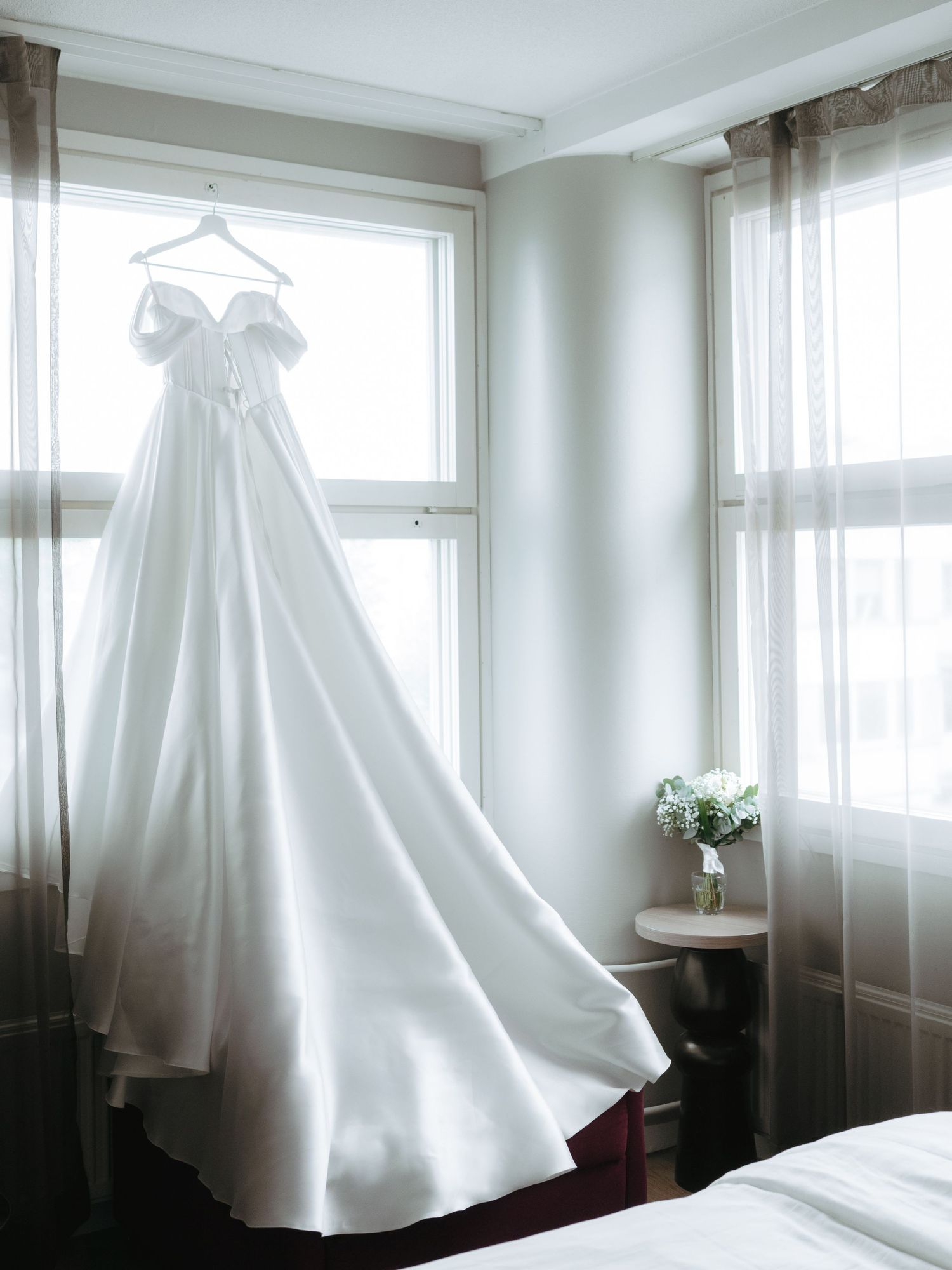 Elegant white off-shoulder wedding gown with a flowing train hanging by a sunlit window.