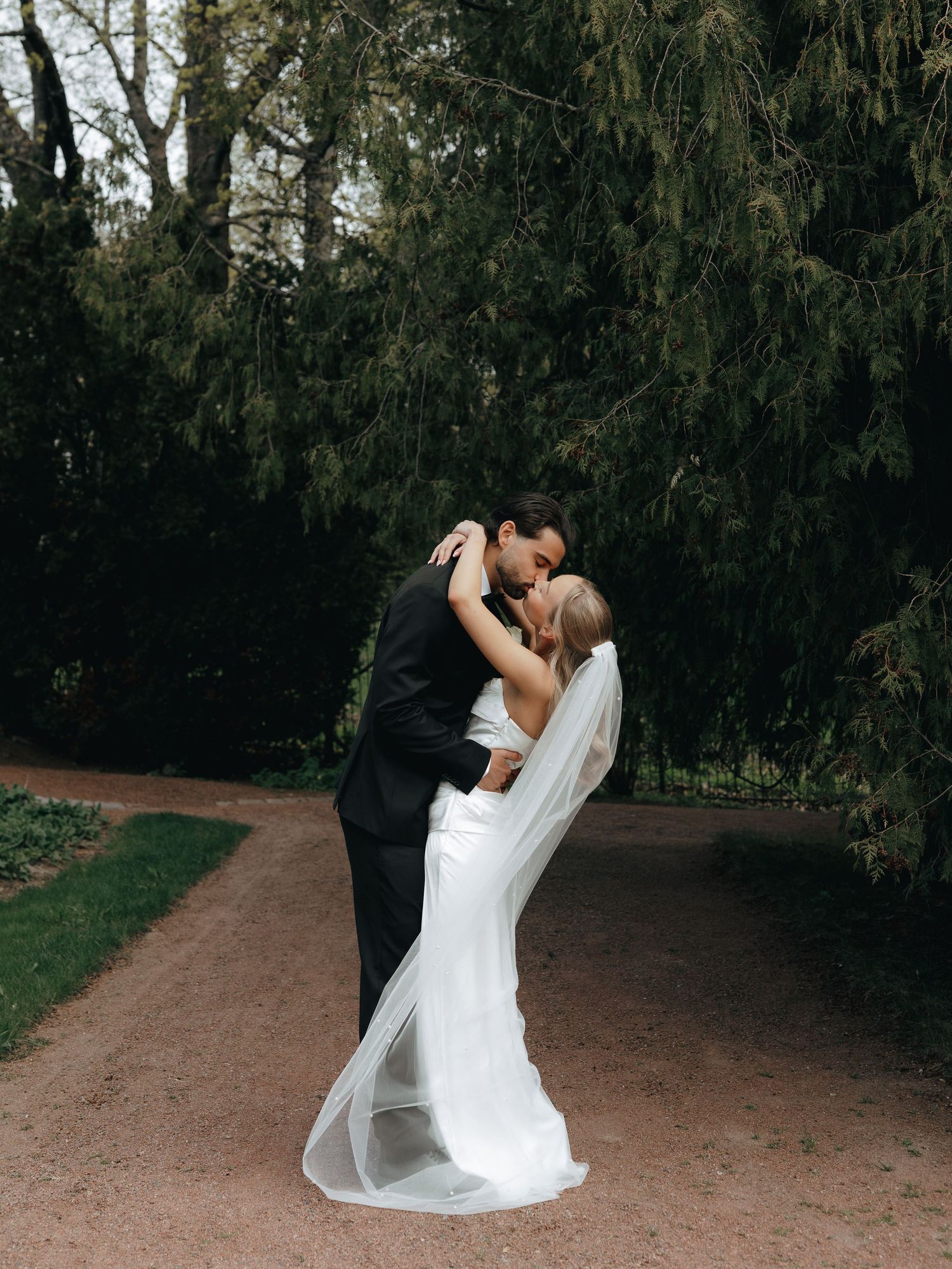 A bride and groom share a romantic kiss on a garden path surrounded by lush green trees on their wedding day.