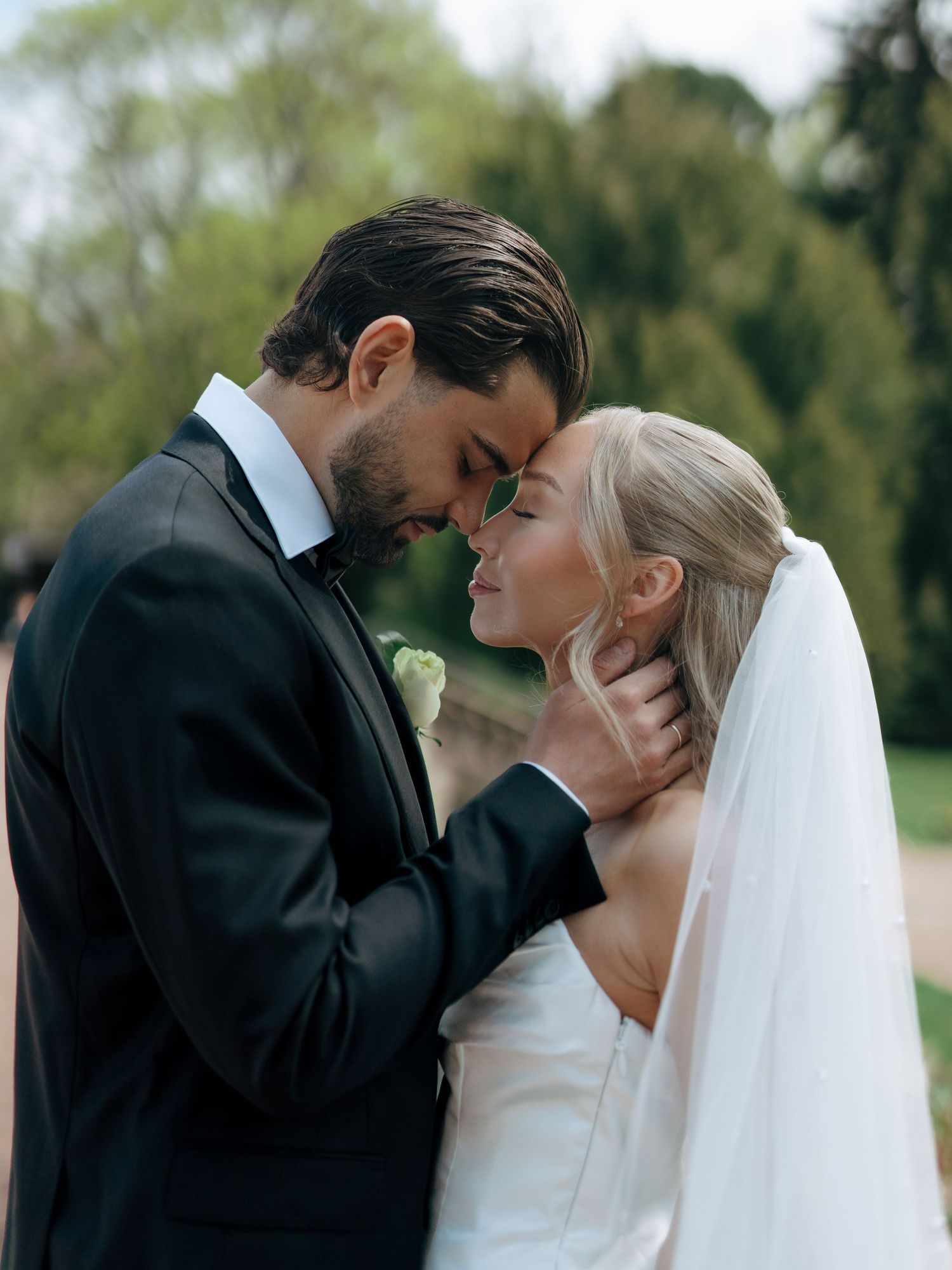 A bride and groom share an intimate forehead touch moment outdoors, wearing a white gown with veil and black tuxedo.