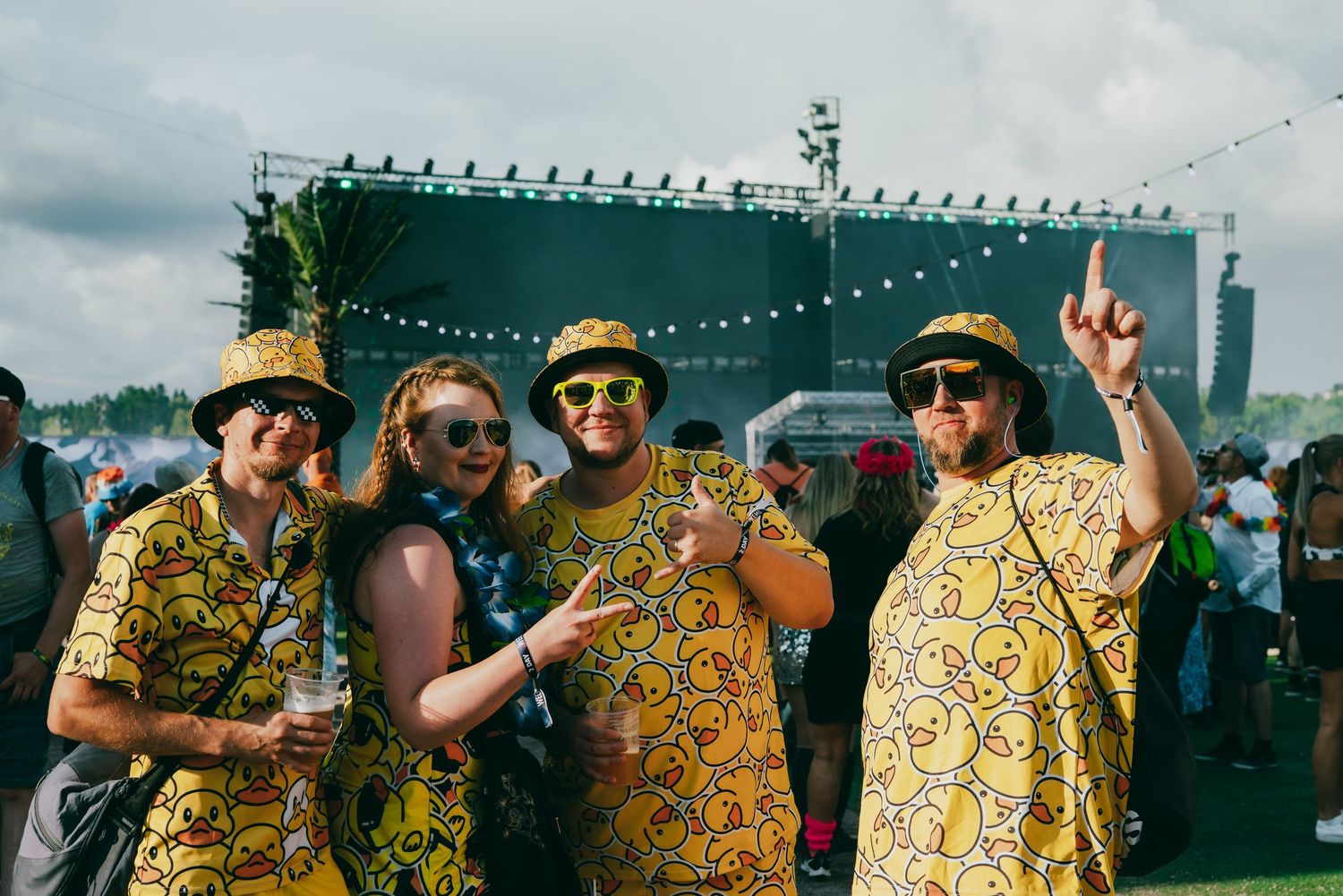 Festival goers in matching yellow patterned outfits pose together at an outdoor music festival with string lights overhead.