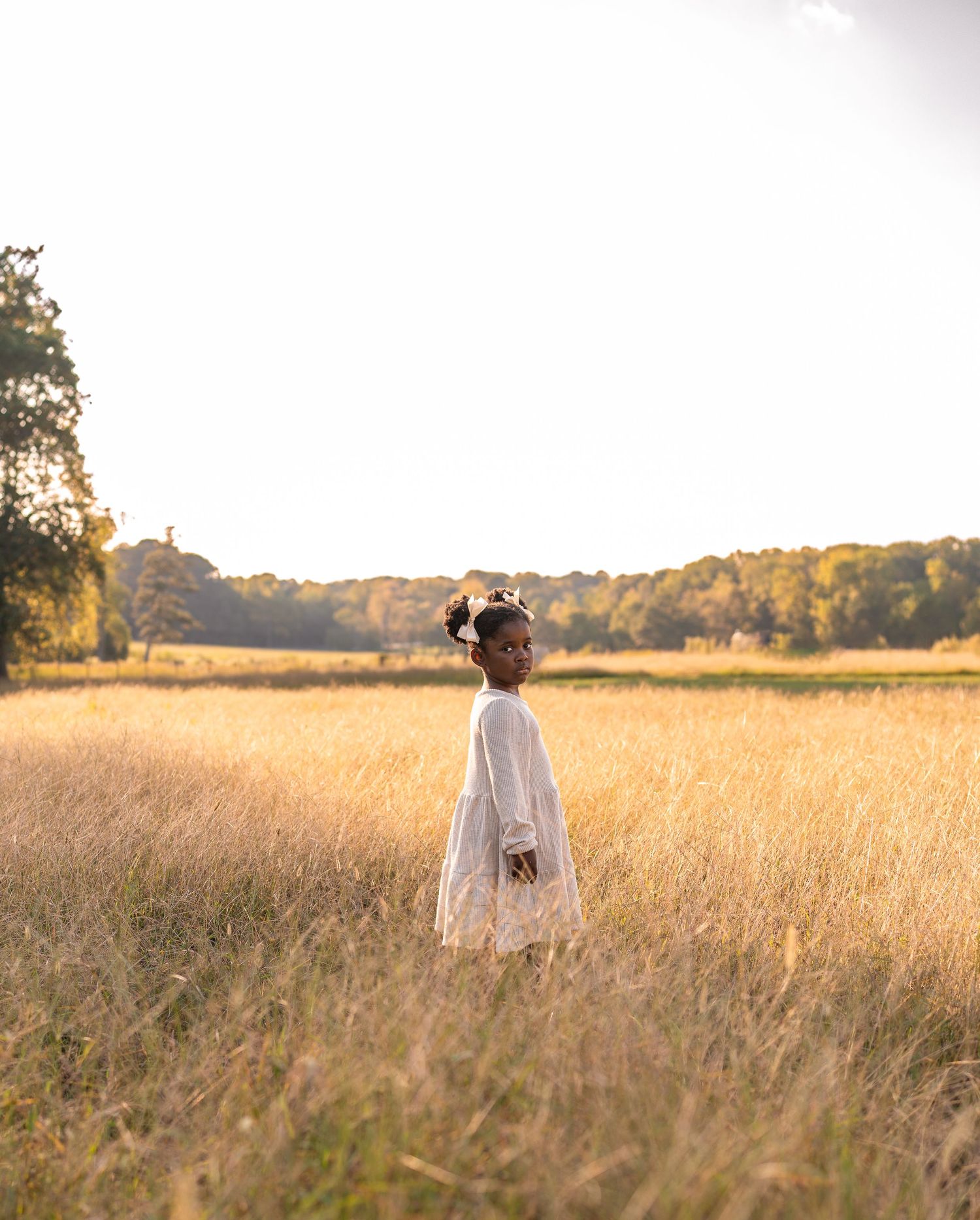 A girl standing in a field during golden hour, with soft, warm sunlight illuminating her as she gazes into the distance, creating a serene a