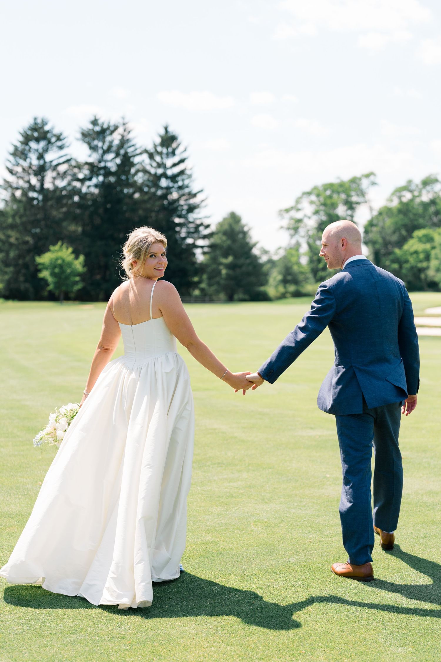 Bride in white gown looks back while holding hands with groom in navy suit walking across green lawn.