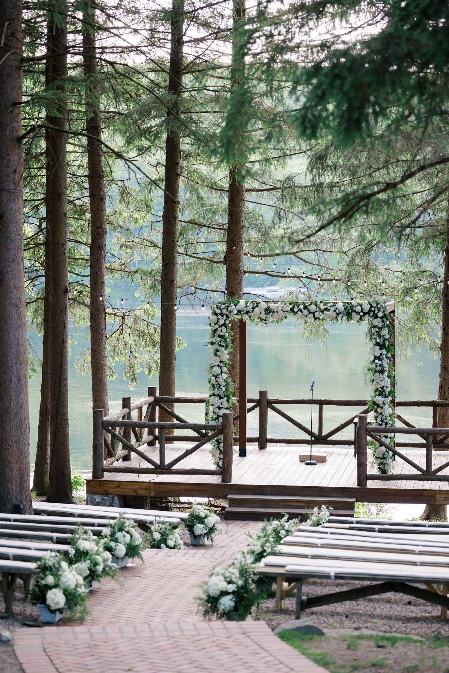 Rustic outdoor wedding ceremony setup with wooden benches and floral arch overlooking a serene lake surrounded by pine trees.