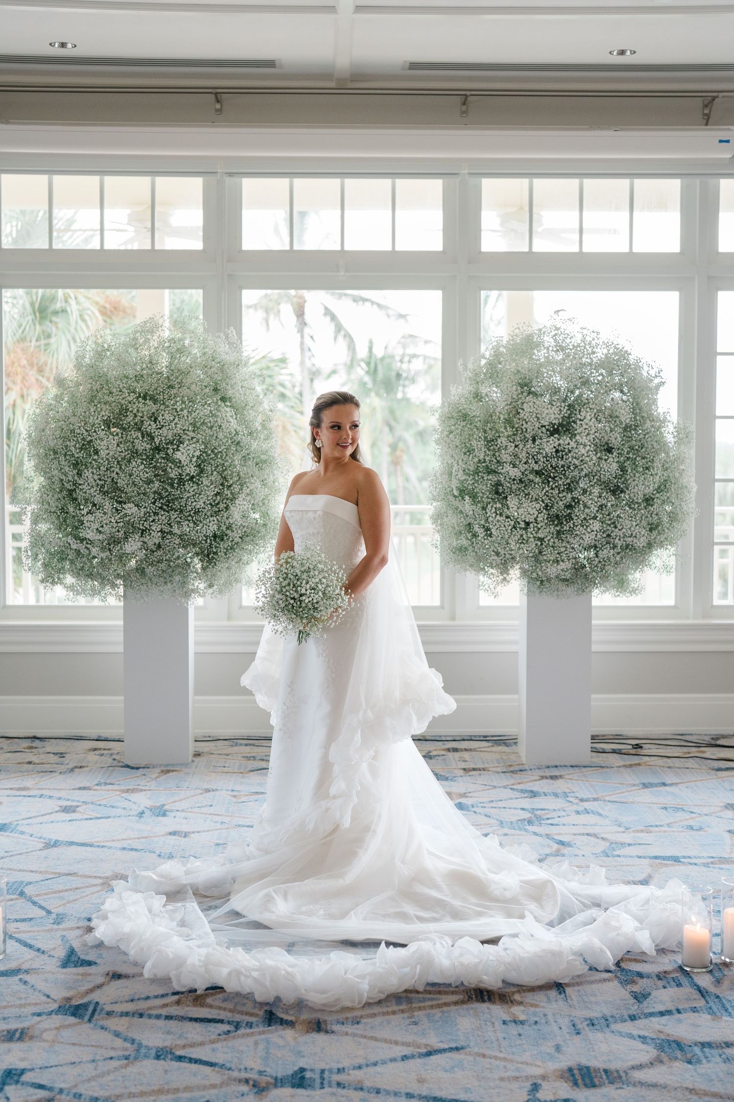 A beautiful wedding dress flows across a patterned carpet in a bright white studio space with baby's breath floral arrangements.