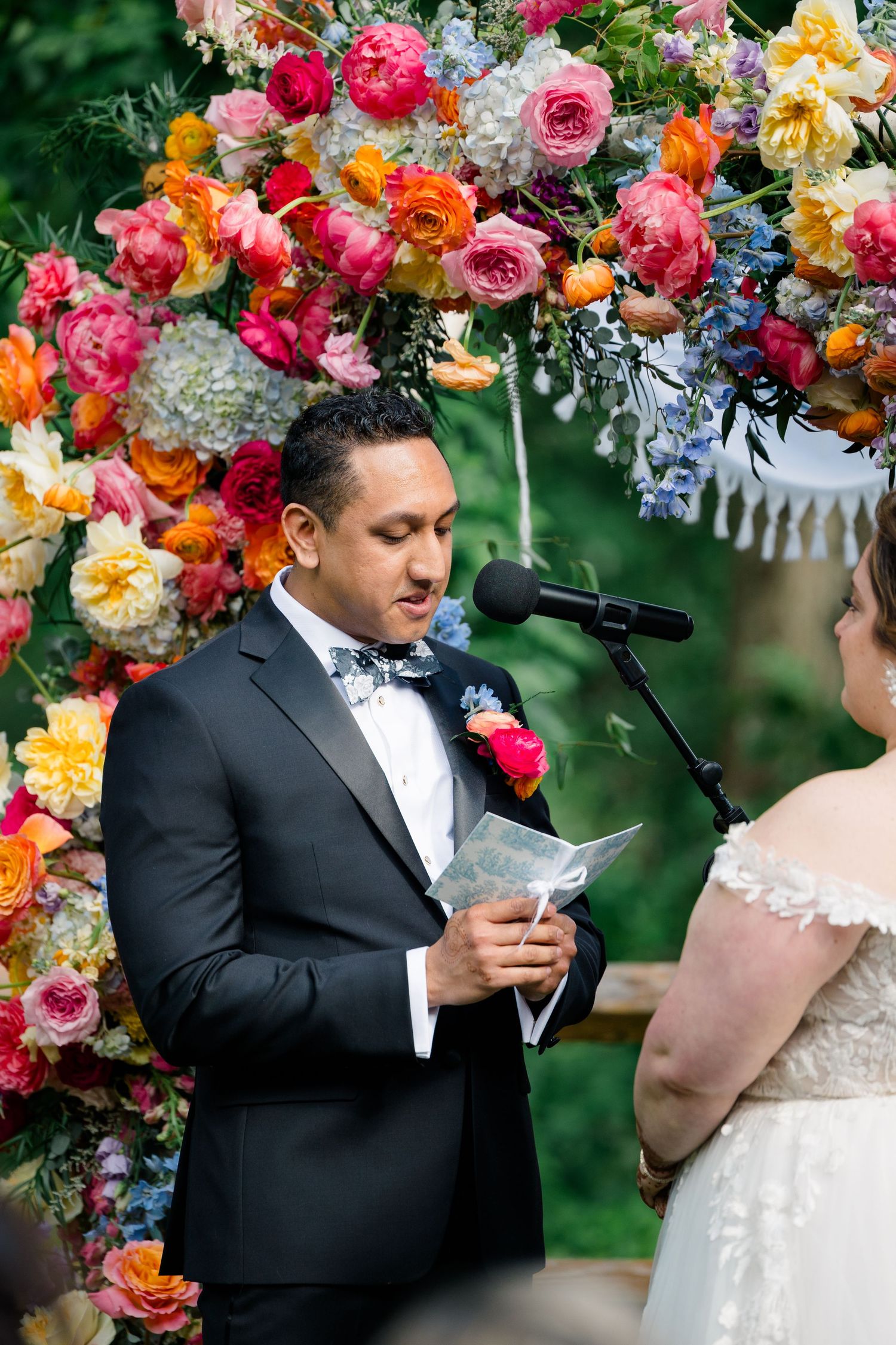 Wedding ceremony under colorful floral arch with officiant reading vows.
