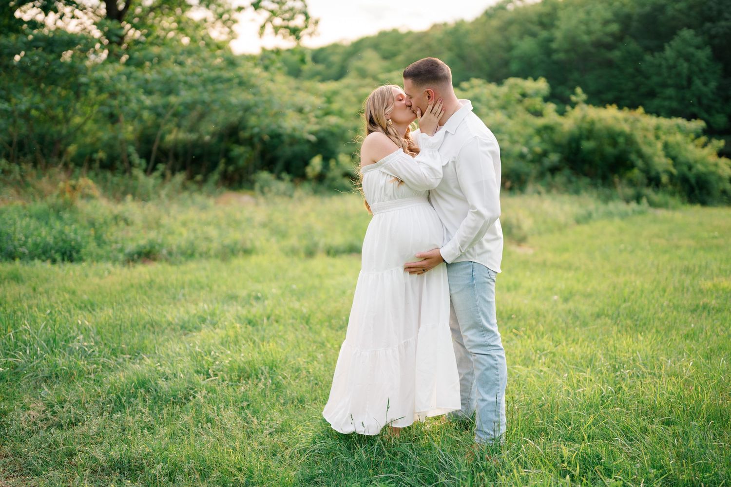 Romantic maternity moment captured in natural meadow setting during sunset.