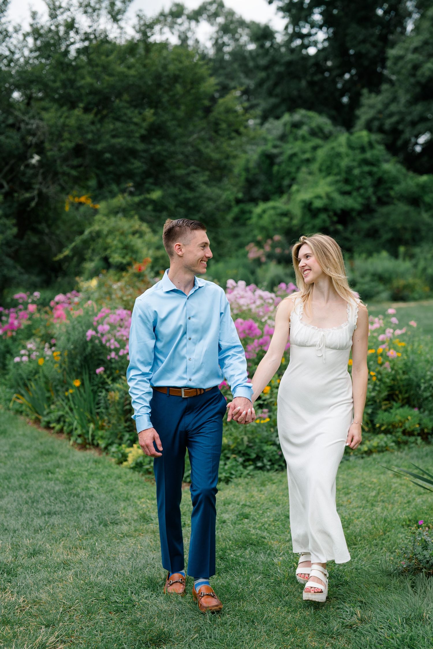 Couple holding hands walking through a lush garden with colorful flowers, man in blue shirt and woman in white dress.