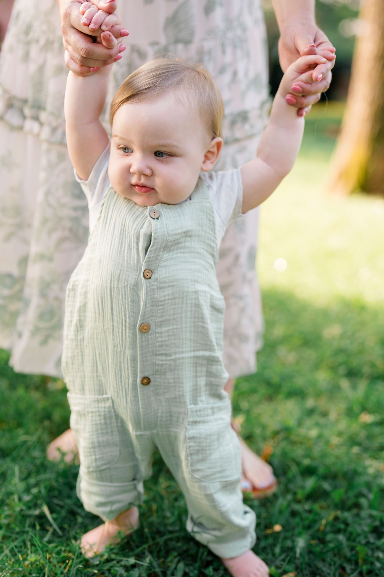A baby in a sage green linen romper takes first steps outdoors, supported by an adult holding both hands on green grass.