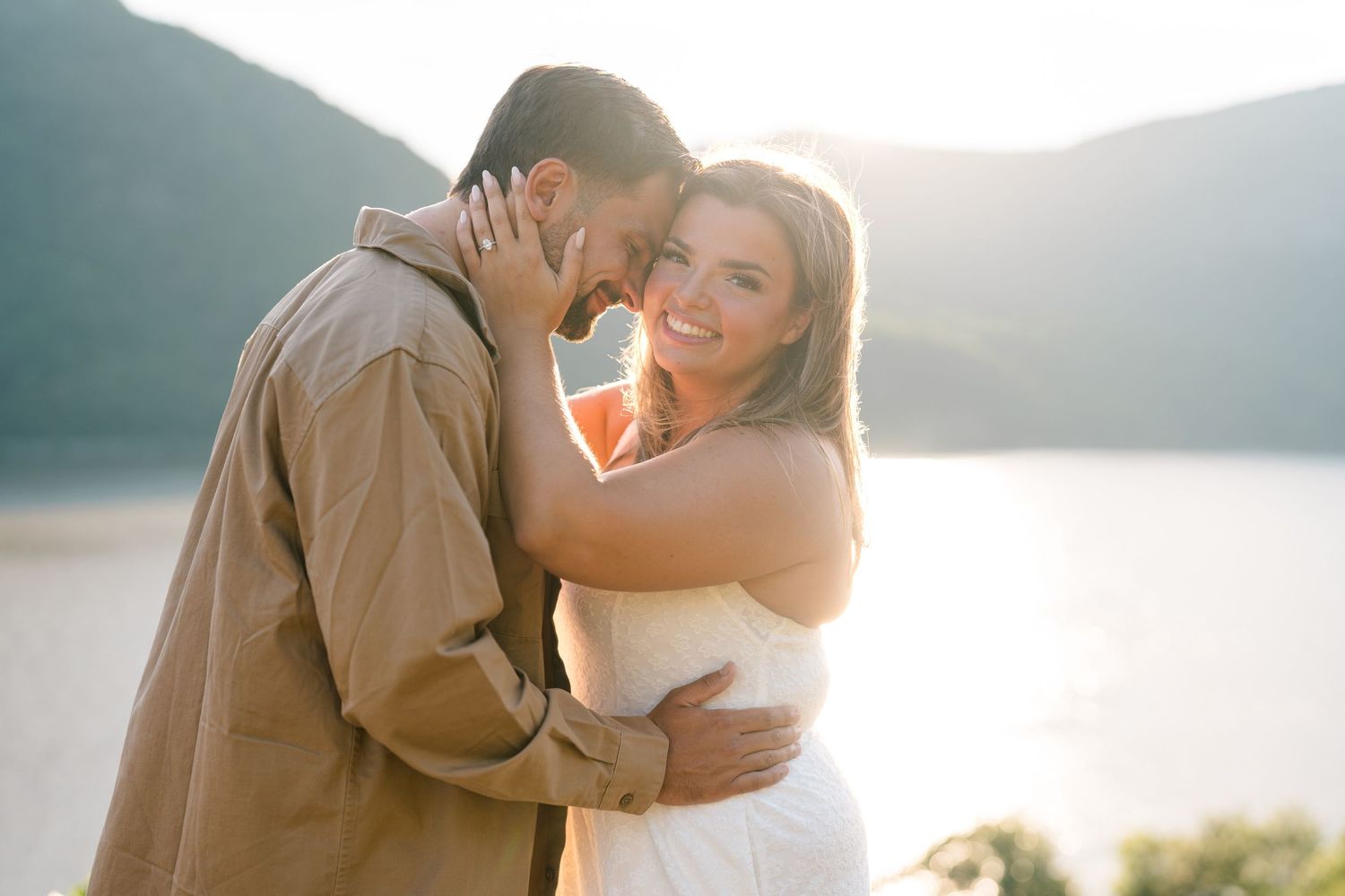 Couple embracing outdoors near a mountain lake at sunset, woman in white dress smiling, man in tan jacket.