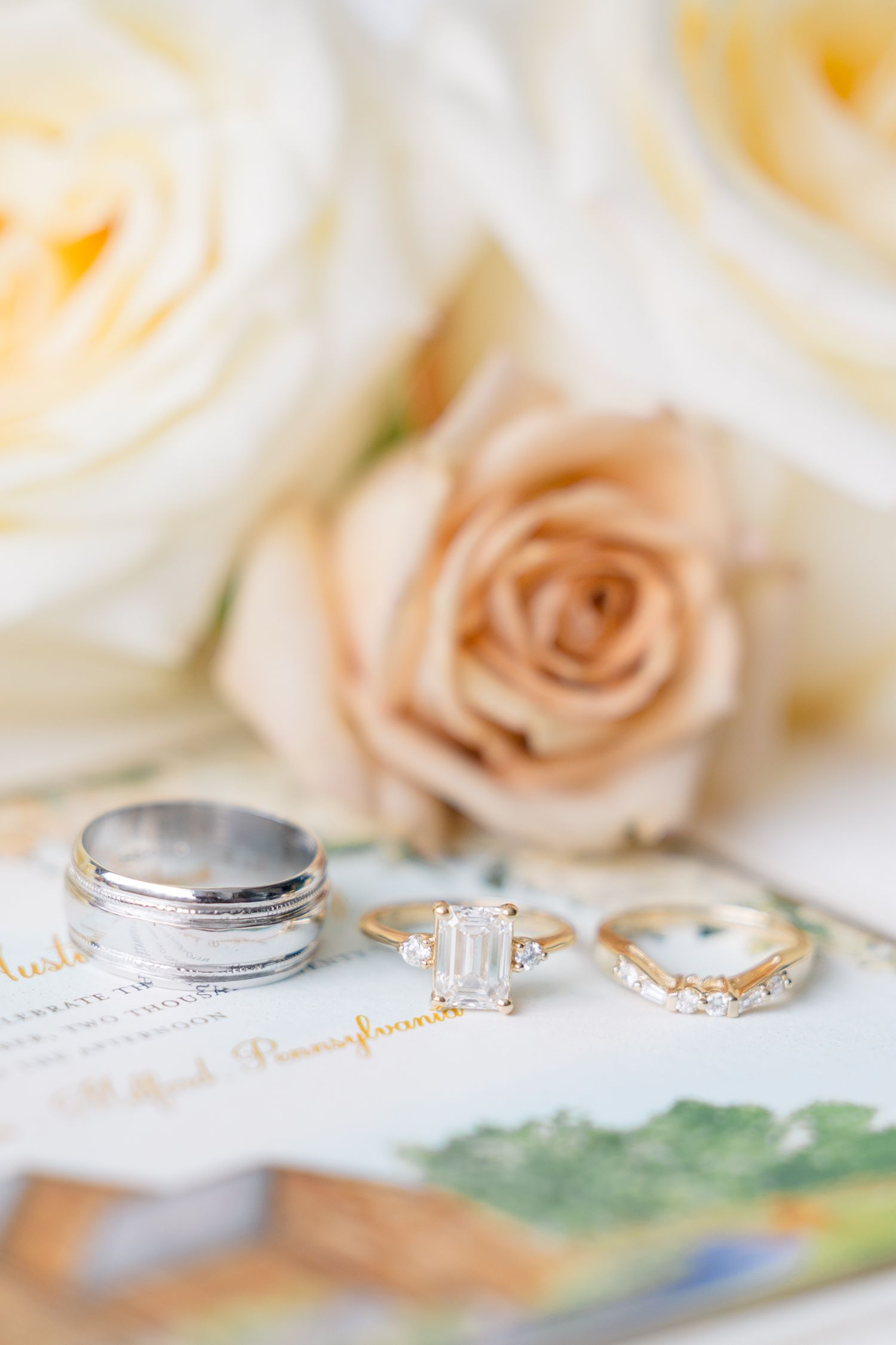Gold engagement ring with emerald-cut diamond and wedding bands displayed on invitation with soft pink and white roses backdrop.