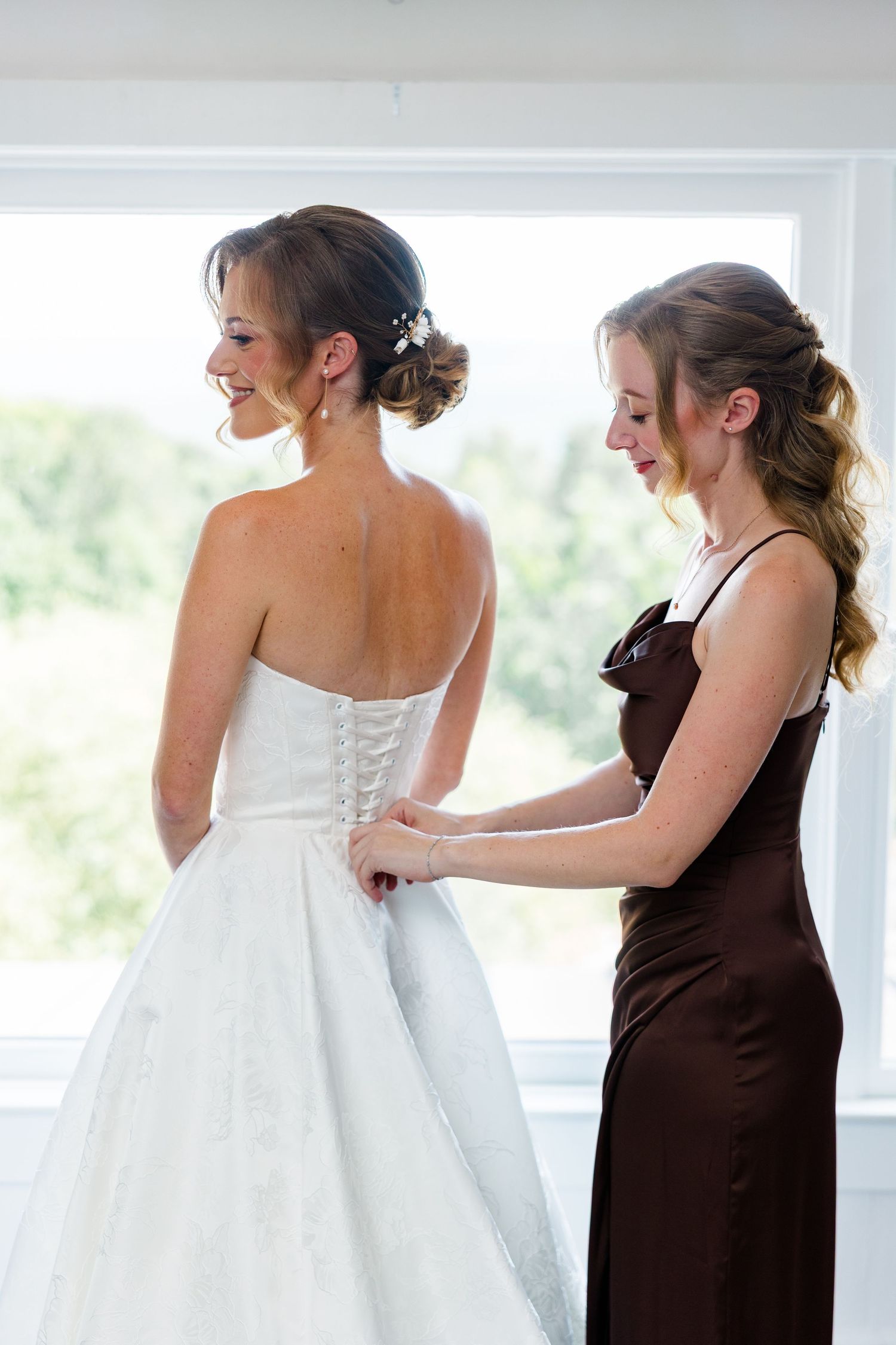 Bridesmaid in brown satin dress laces up the back of a smiling bride's white strapless wedding gown.