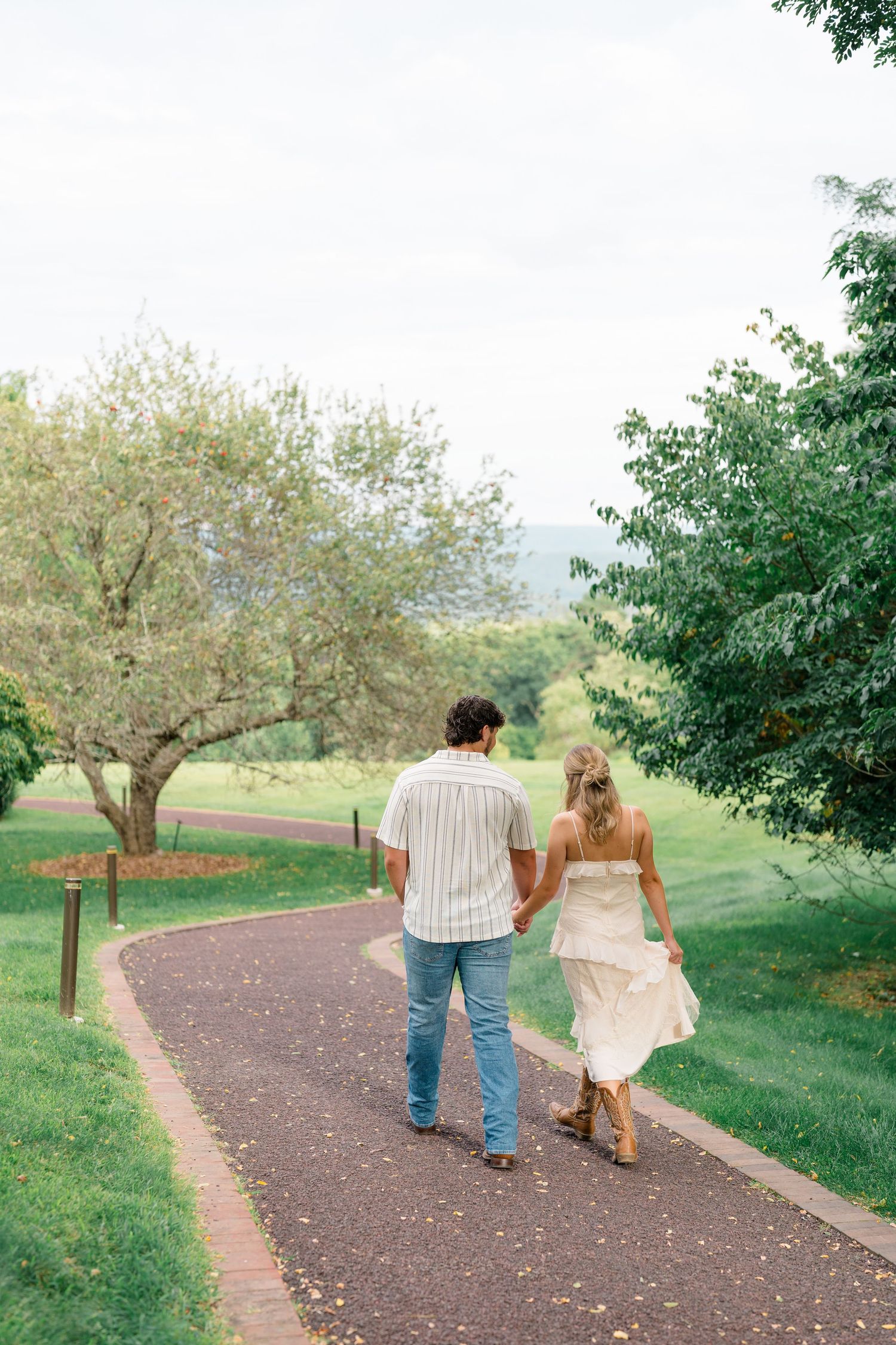 Couple holding hands walking down a scenic garden path surrounded by lush green trees.