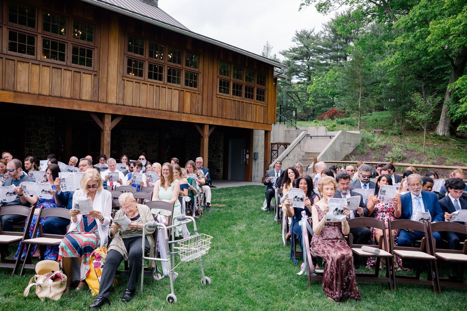 Wedding guests seated on white chairs outside a rustic wooden venue surrounded by green trees.