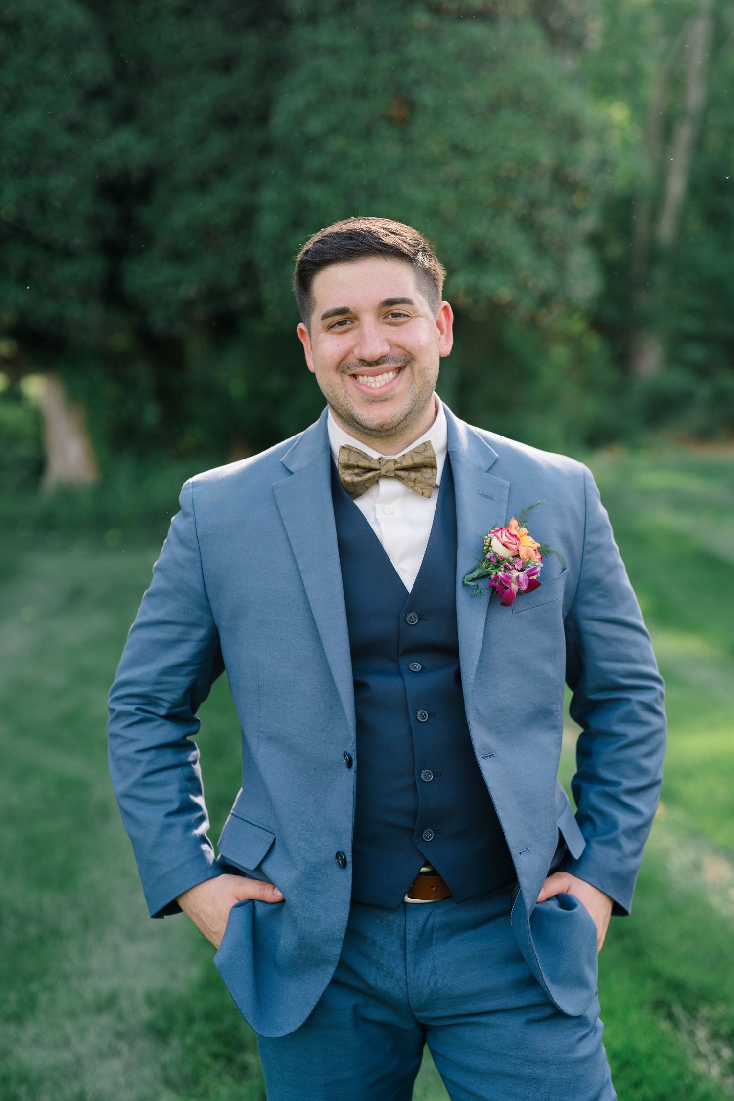 Groom in blue three-piece suit and bow tie smiles against greenery backdrop.