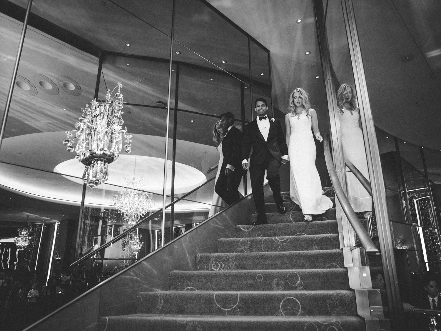 A glamorous couple descends an elegant staircase beneath a sparkling crystal chandelier in a black and white photograph.
