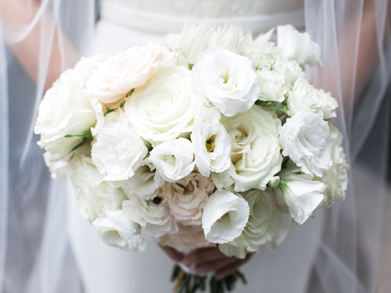 An elegant bridal bouquet of pure white roses and lisianthus flowers wrapped with a delicate veil in soft focus.
