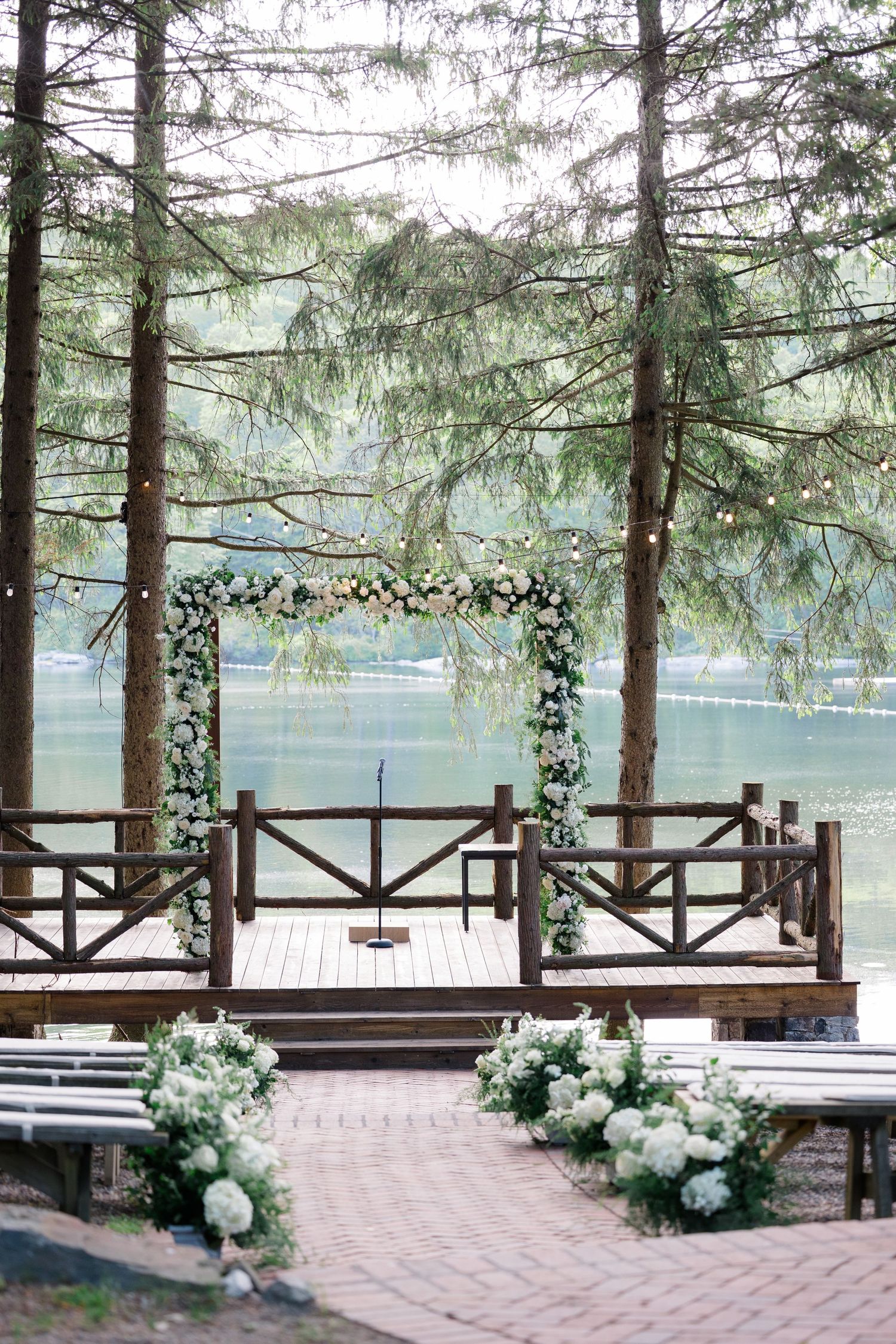 A romantic lakeside wedding ceremony setup with a floral arch and wooden benches framed by tall evergreen trees.