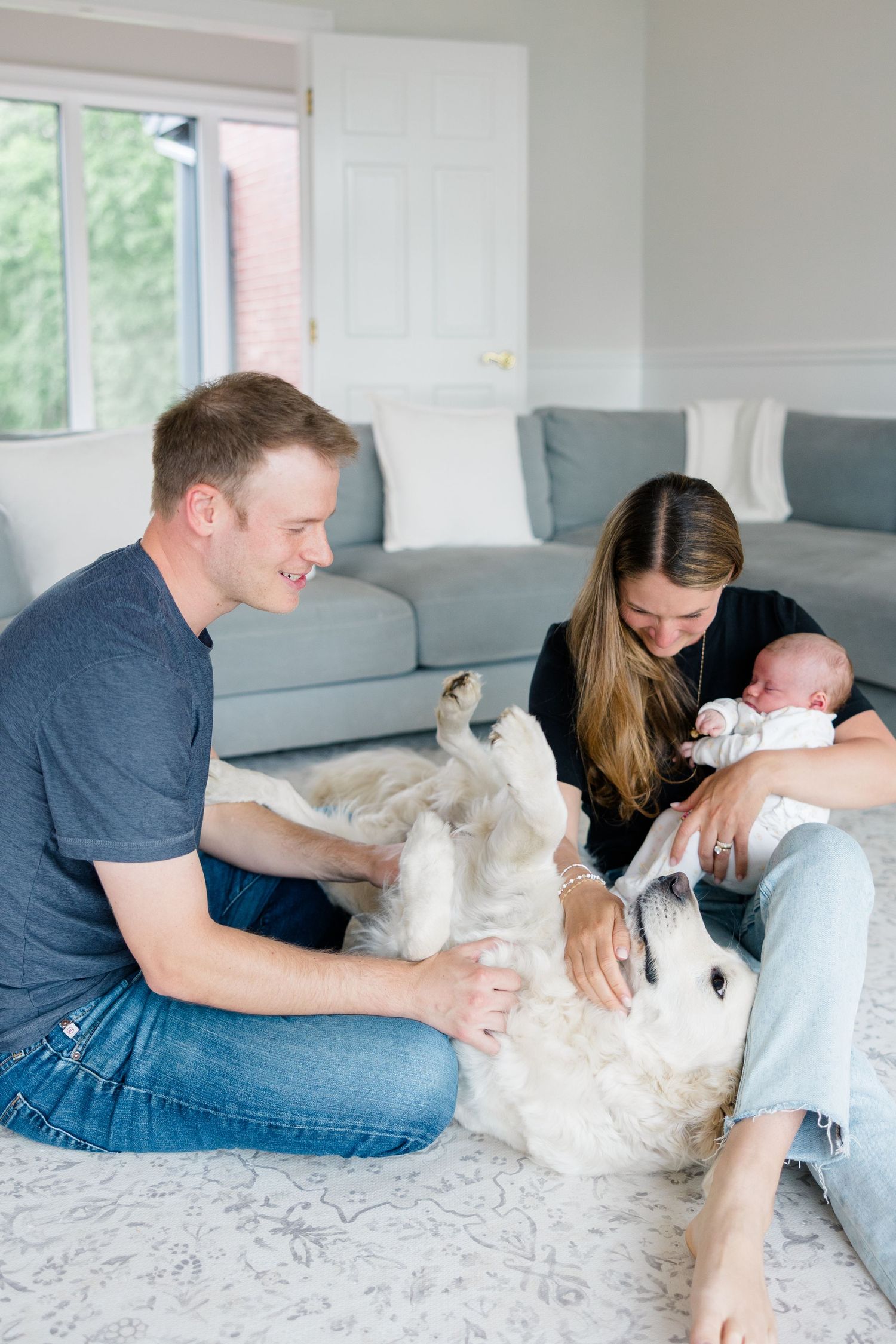 Family relaxing on carpet with golden retriever while holding newborn.