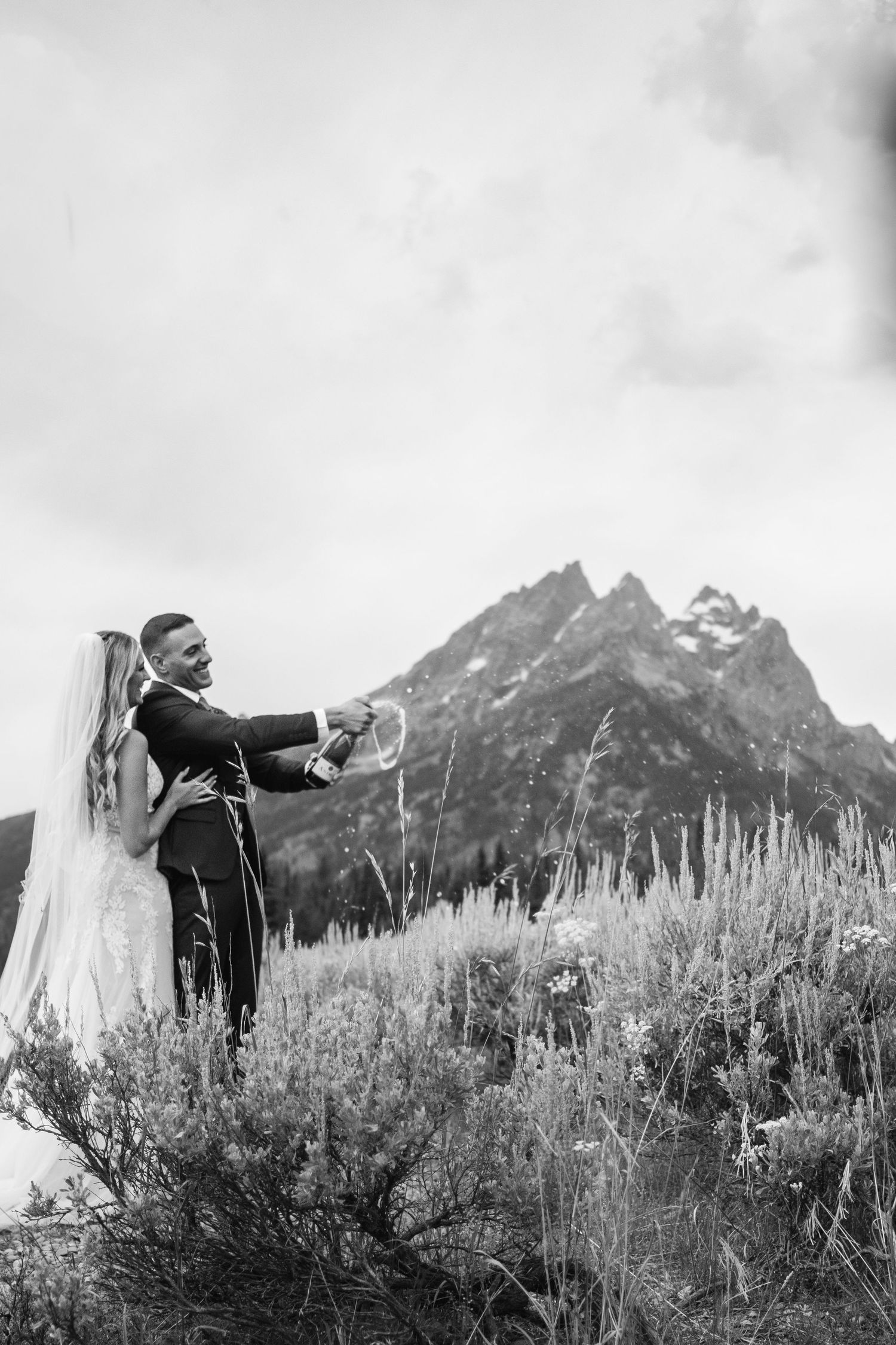 Silhouetted figures share an intimate embrace against dramatic mountain peaks and tall wildgrasses in black and white.