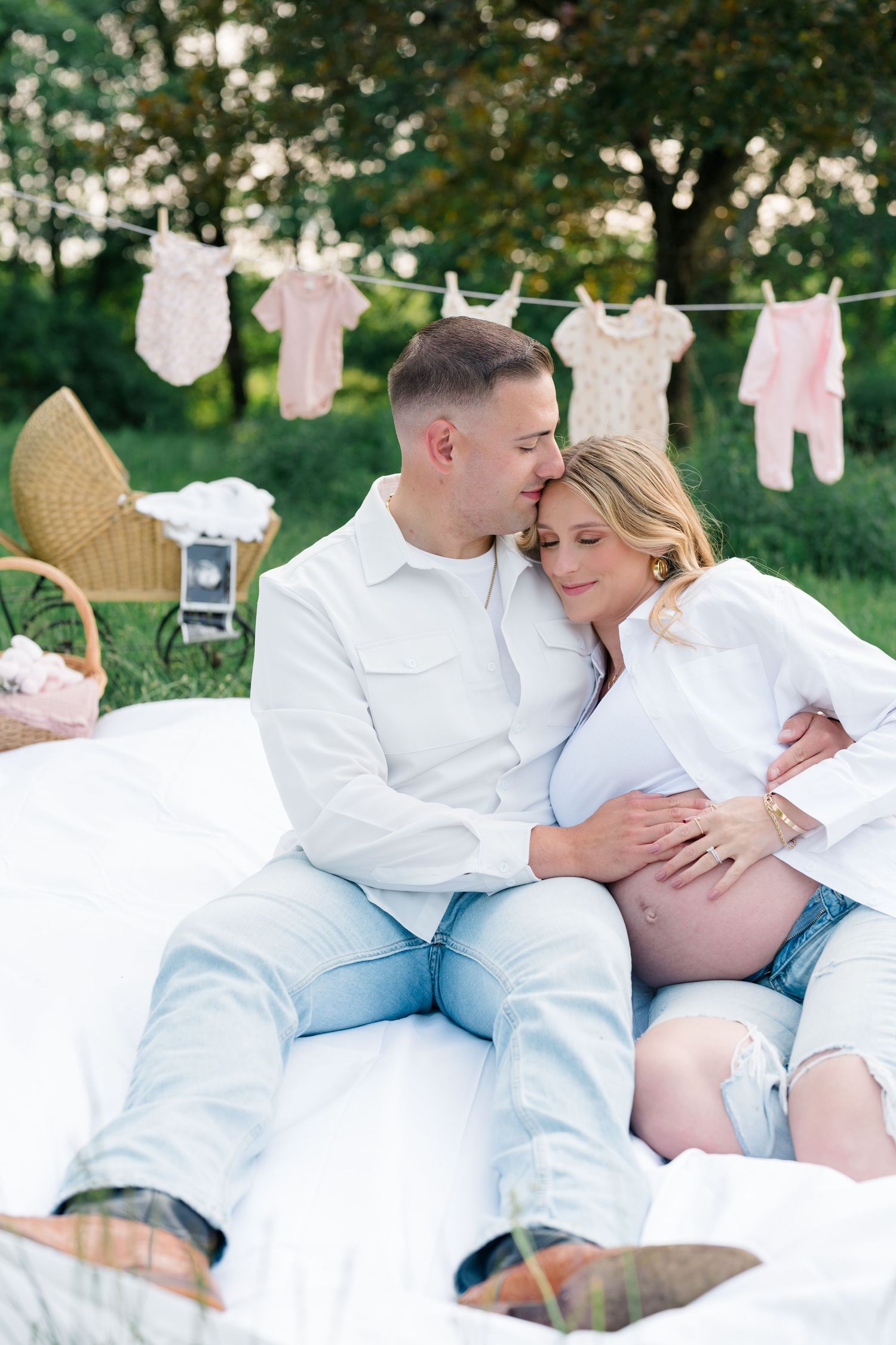Expecting couple poses by clothesline with baby outfits during outdoor maternity photoshoot.