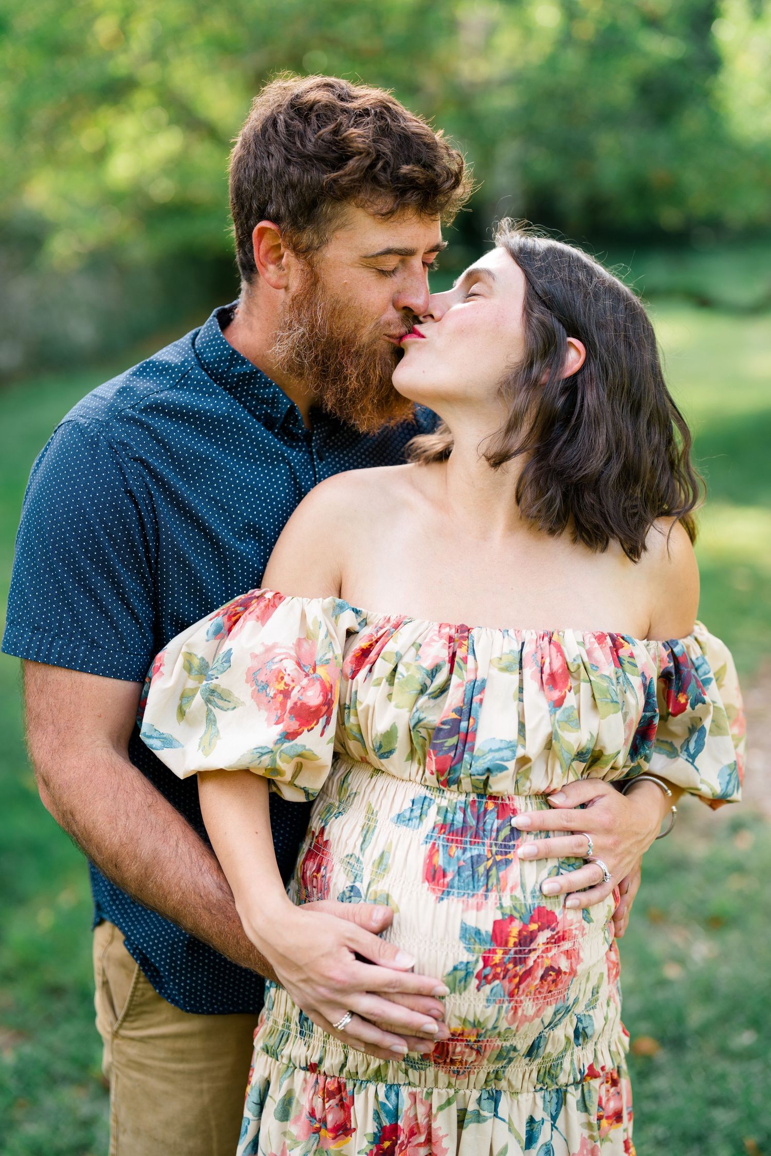 Couple sharing a kiss outdoors, man embracing pregnant woman in floral off-shoulder dress in a lush green garden.