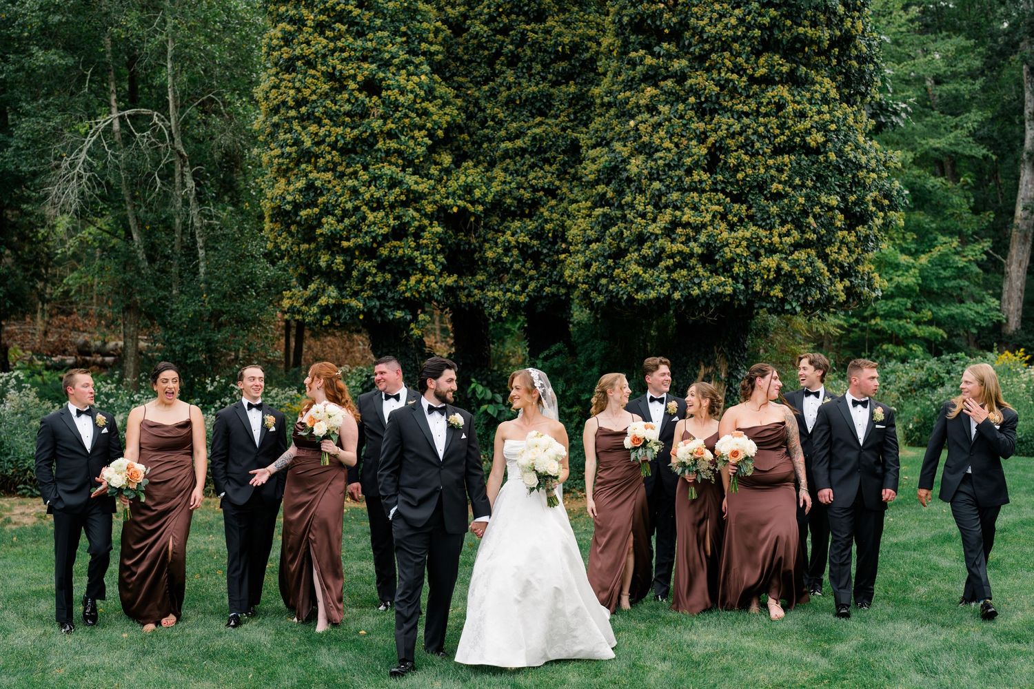 Wedding party in black tuxedos and brown gowns poses with bride and groom on lush green lawn.