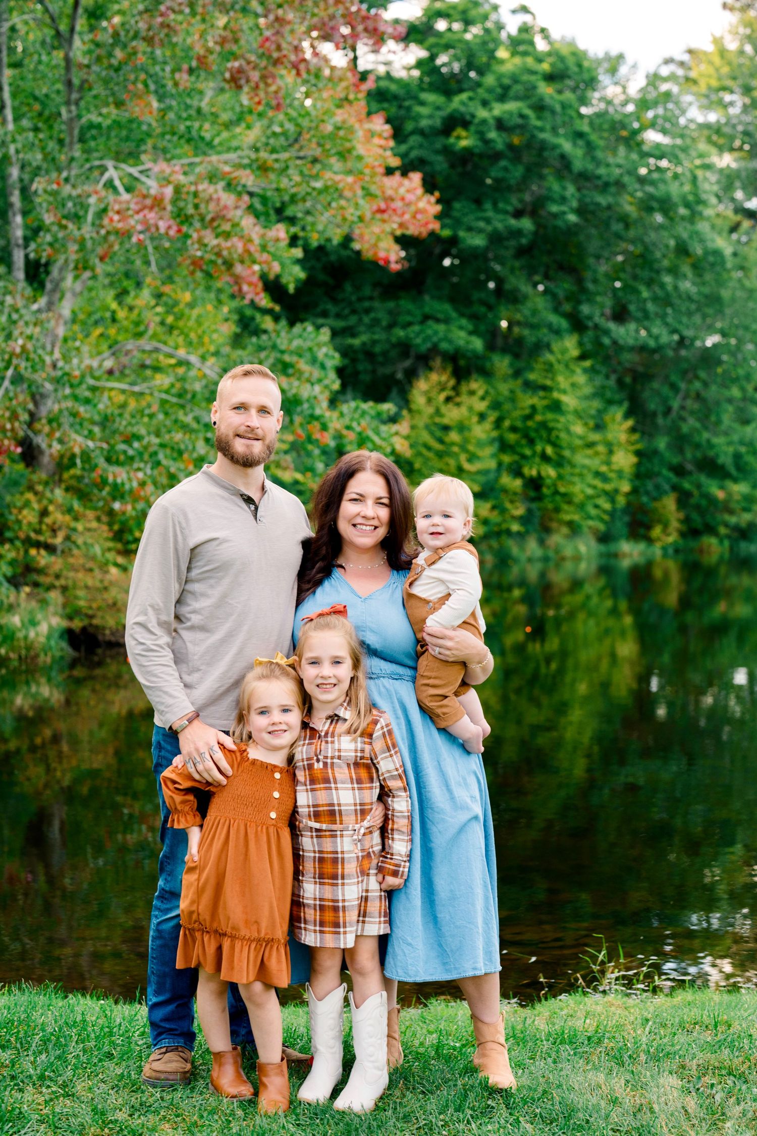 Family of five posing outdoors near a pond with lush green trees in the background during fall season.