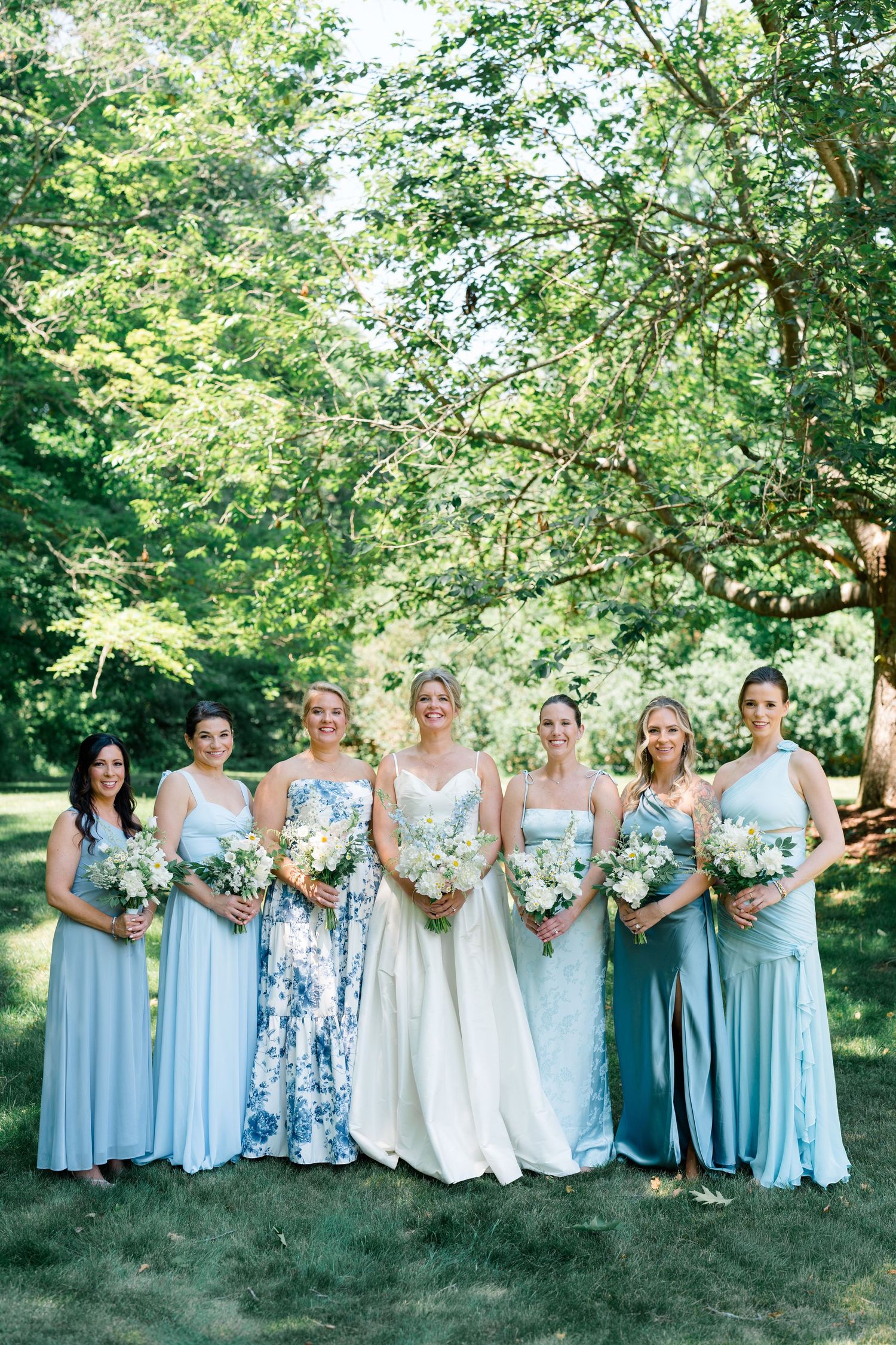Bride in white gown poses with six bridesmaids in blue dresses holding white bouquets outdoors.
