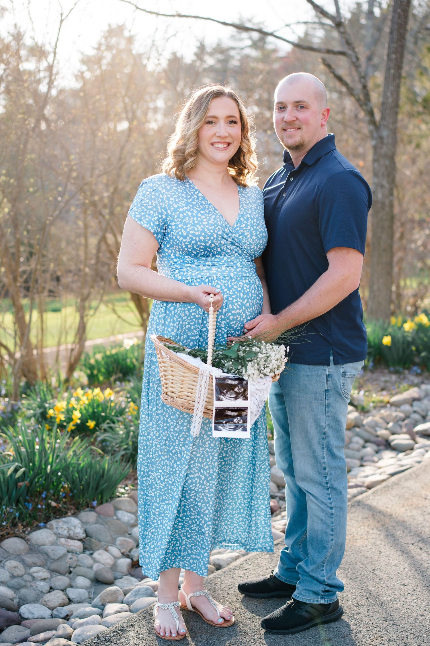 A couple stands outdoors on a stone path holding an ultrasound photo and basket with yellow daffodils blooming nearby.