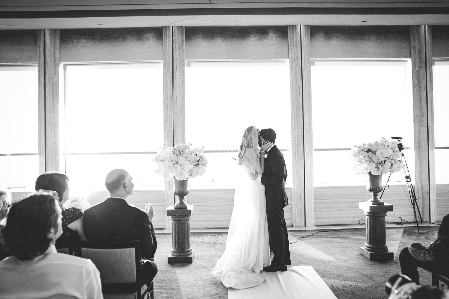 Intimate wedding ceremony in bright room with large windows and floral arrangements.