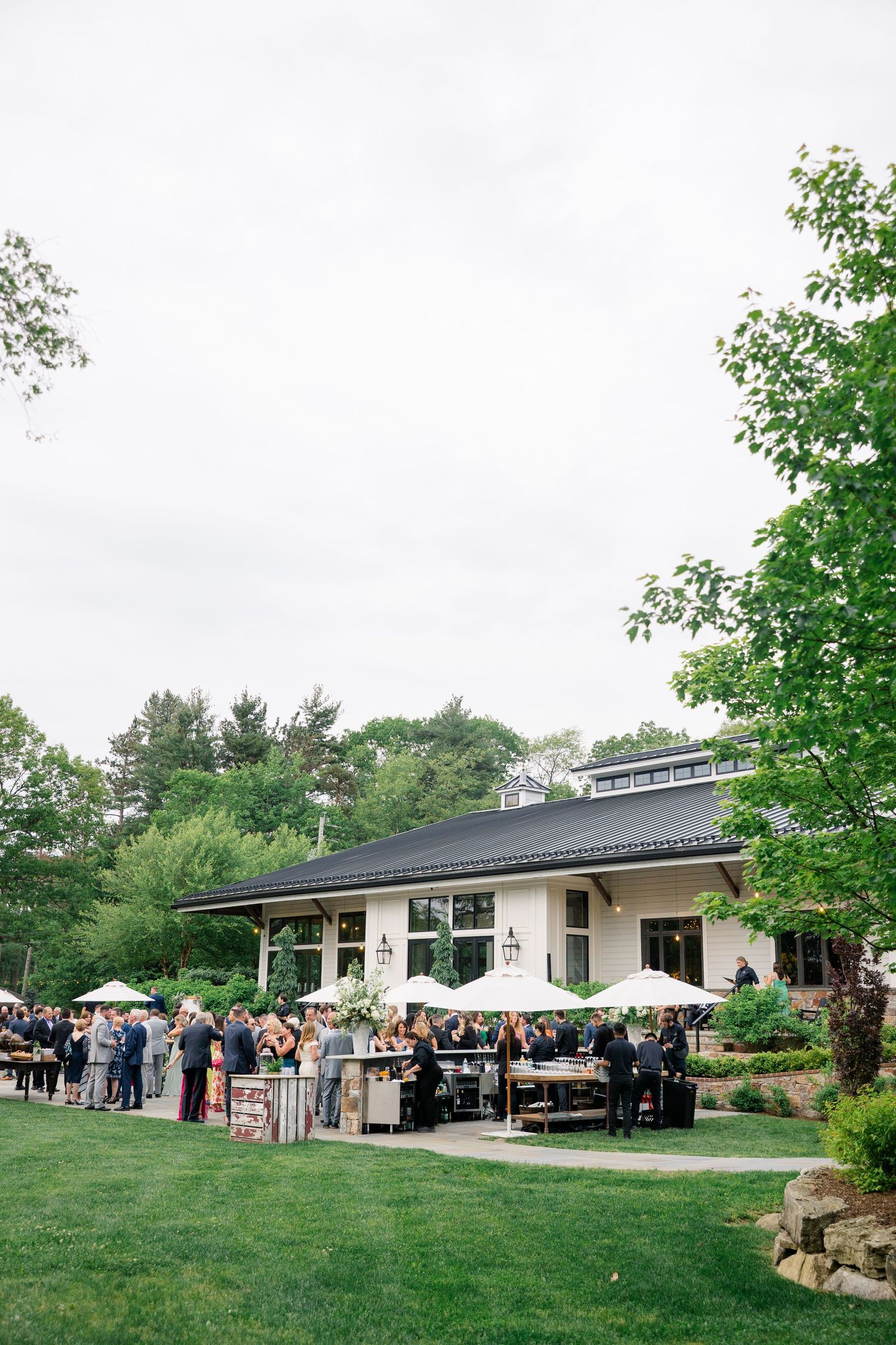 Outdoor wedding reception venue with guests gathered on lawn in front of a modern rustic building.