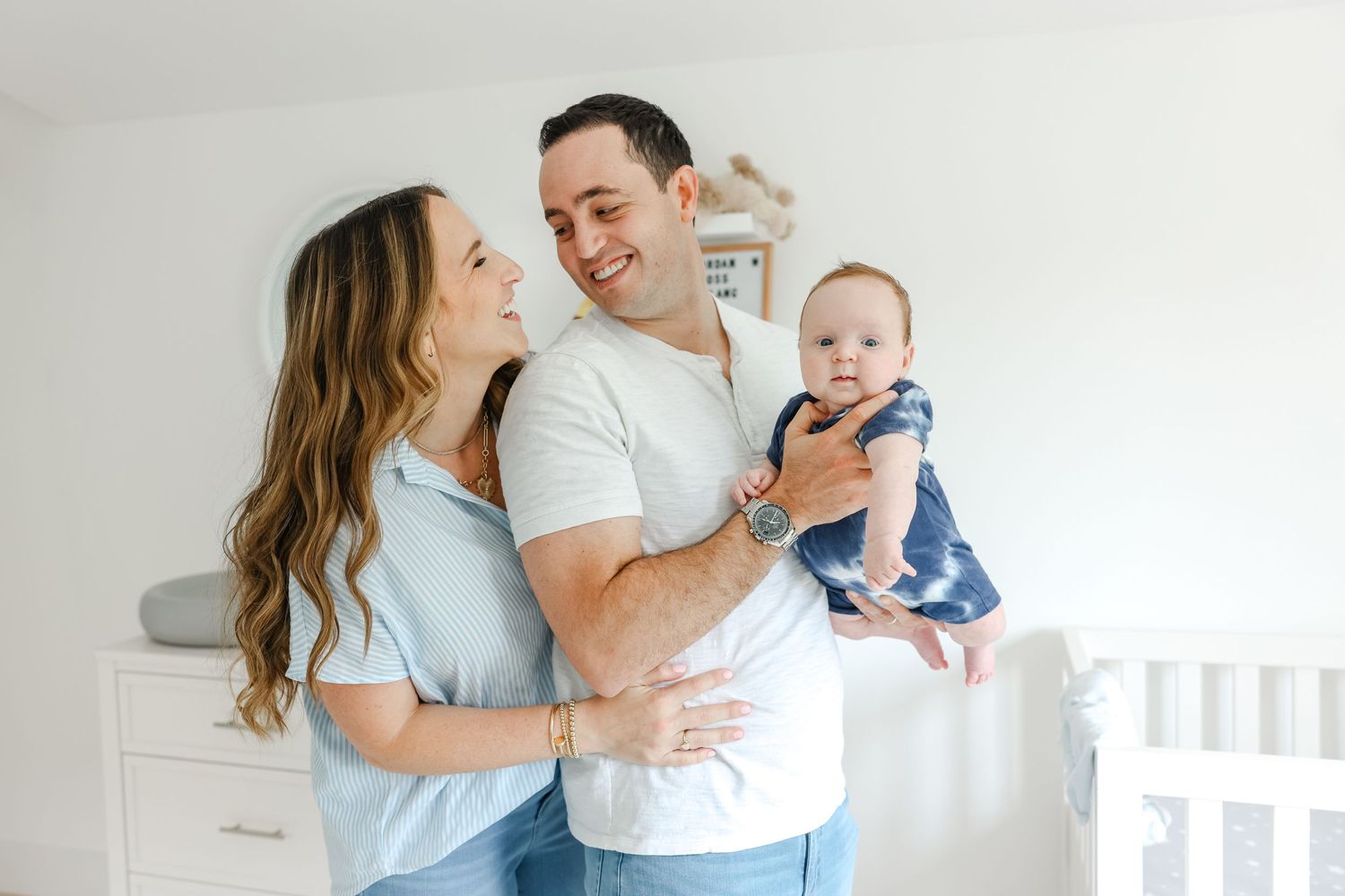 A couple wearing light blue and white smiles while holding their baby in a bright white nursery.