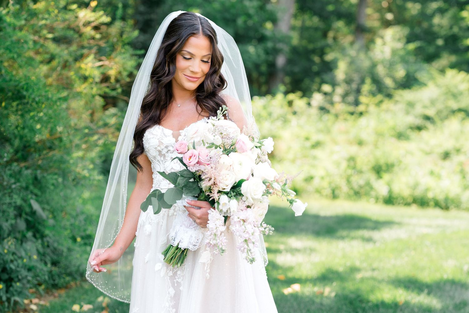 A bride in a white lace gown holds a soft pink and white floral bouquet while standing in a sunlit garden.