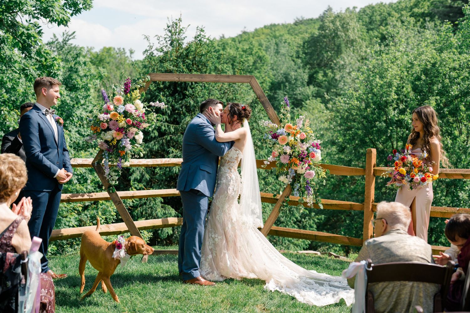 A couple exchanges vows during an outdoor ceremony on a wooden deck surrounded by floral arrangements and greenery.