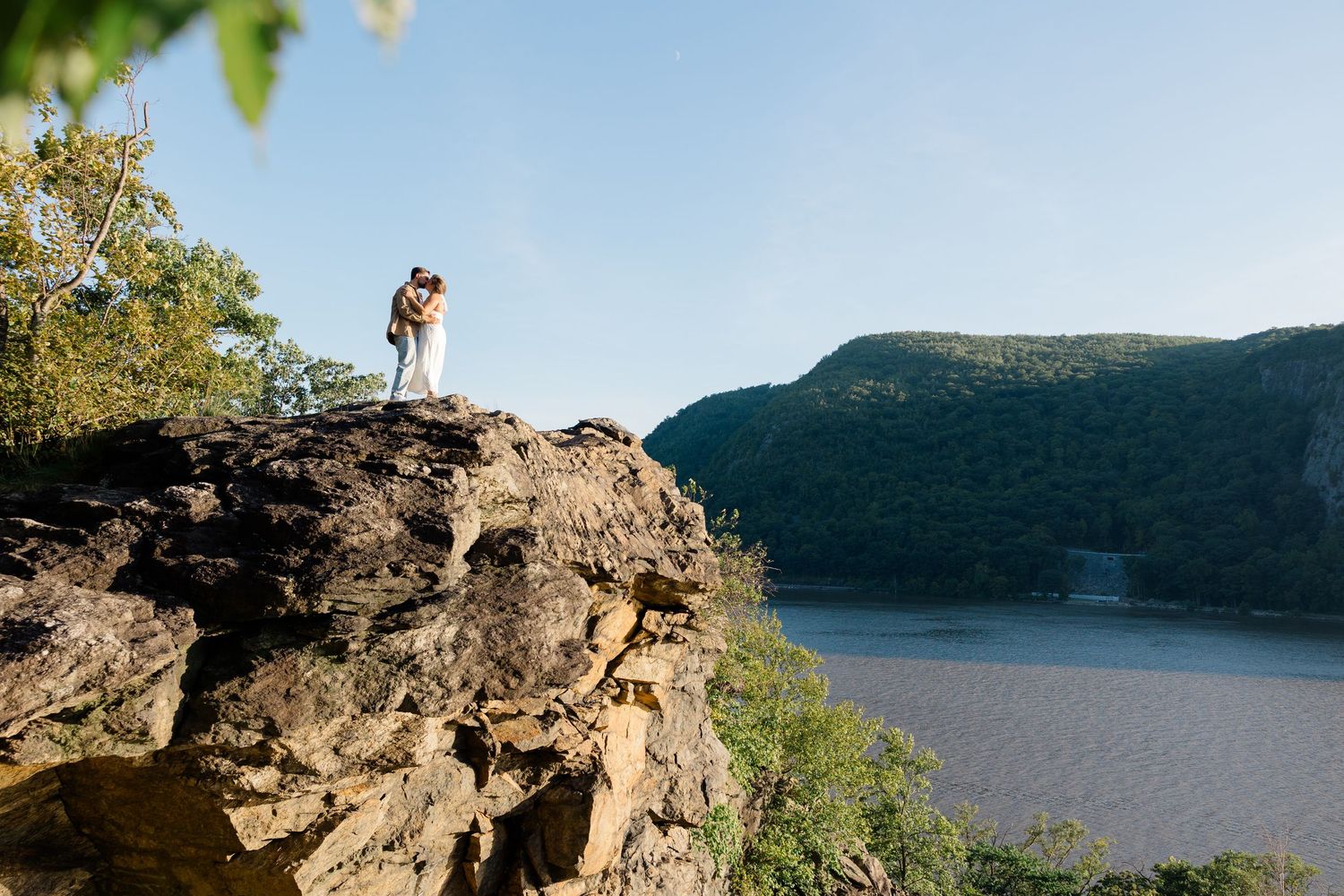 Couple kissing on rocky cliff overlooking scenic river and green mountains under clear blue sky.