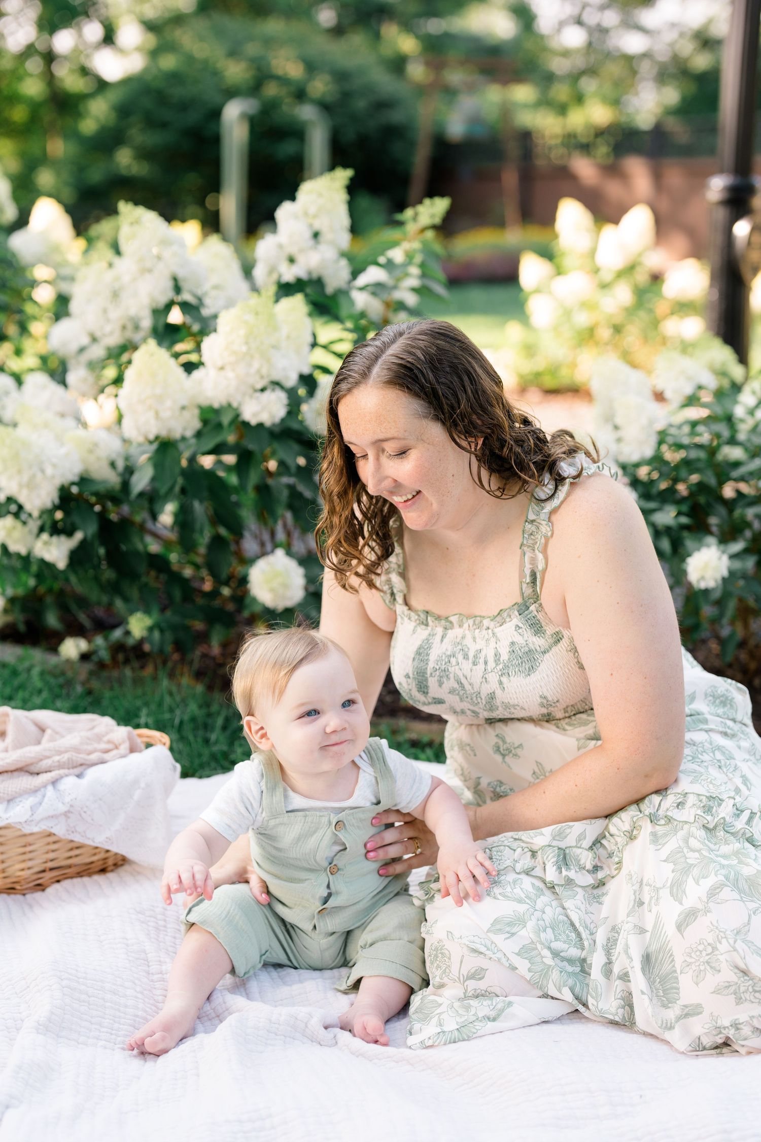 Mother and baby boy sitting on white blanket near white hydrangea flowers in garden.