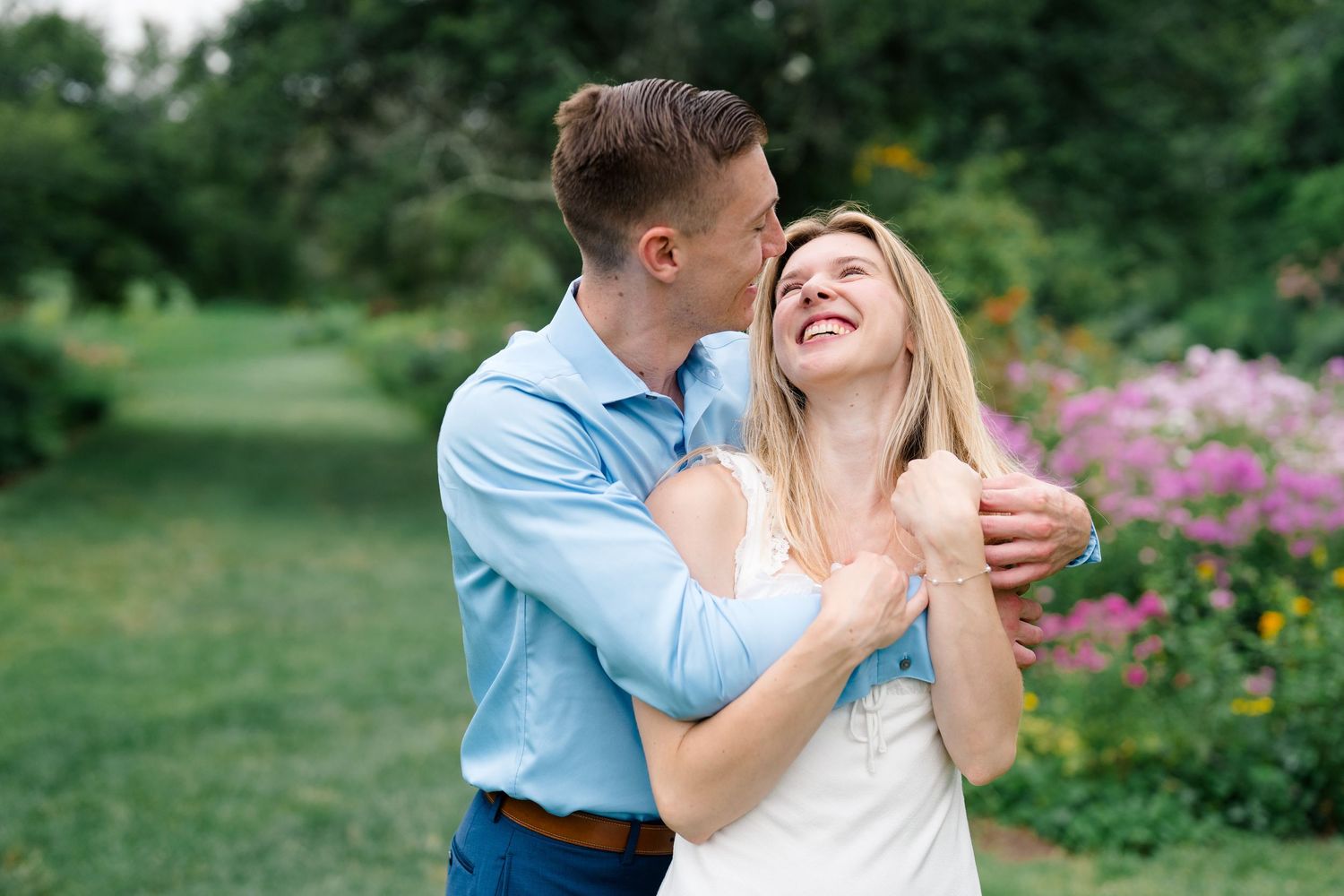 A happy couple embraces outdoors, man in blue shirt kisses smiling blonde woman in white dress amid colorful garden blooms.