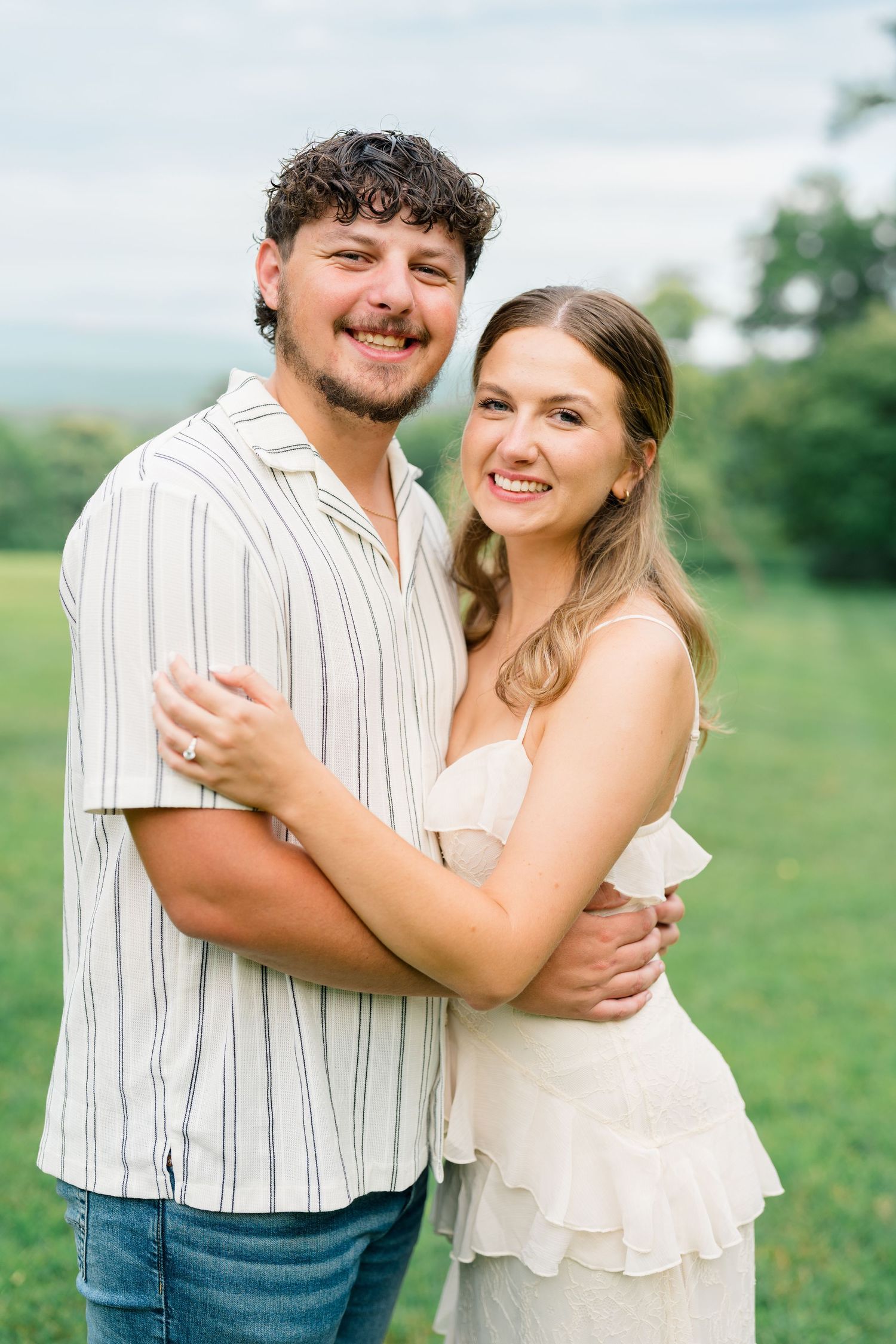 Smiling couple embracing outdoors in a green field, man in striped shirt and woman in white ruffled dress.