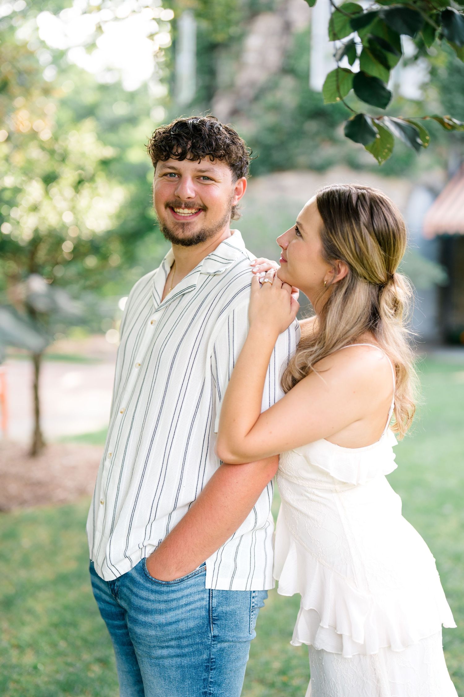 A smiling couple poses outdoors, man in striped shirt and jeans, woman in white dress embracing him from behind.