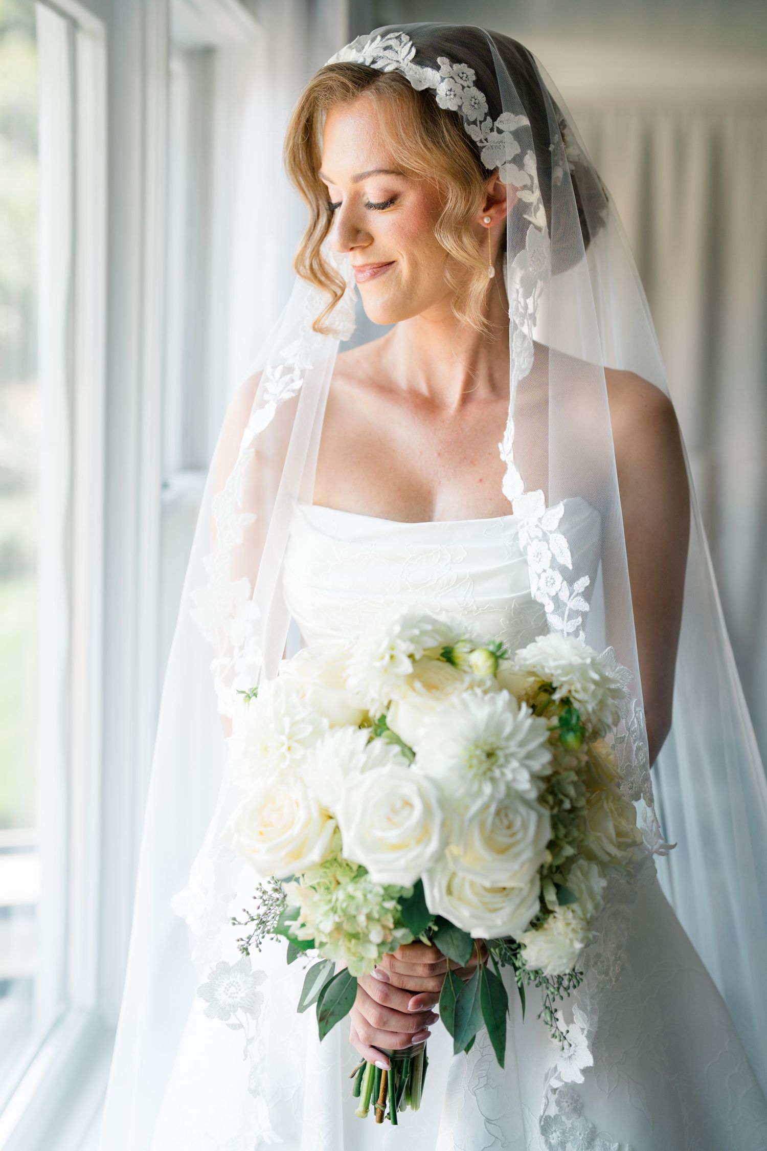 Elegant bride in white gown and lace veil holds a bouquet of white roses and dahlias, gazing down by a window.