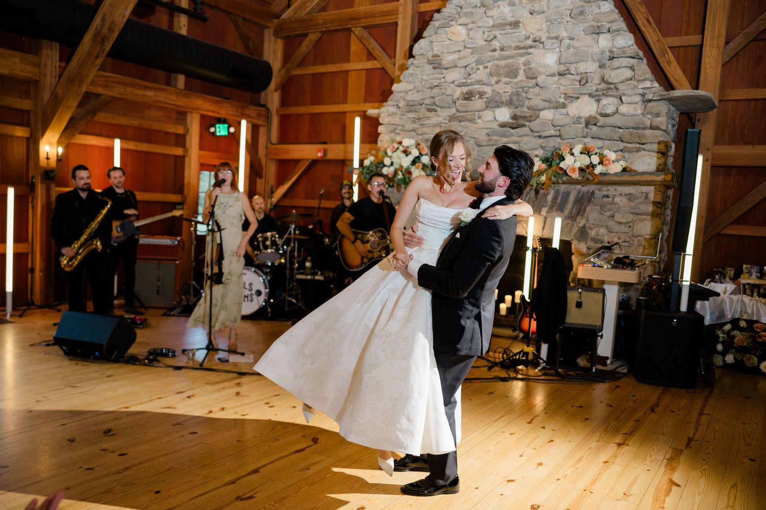 Bride and groom share first dance in rustic barn venue with live band and stone fireplace backdrop.