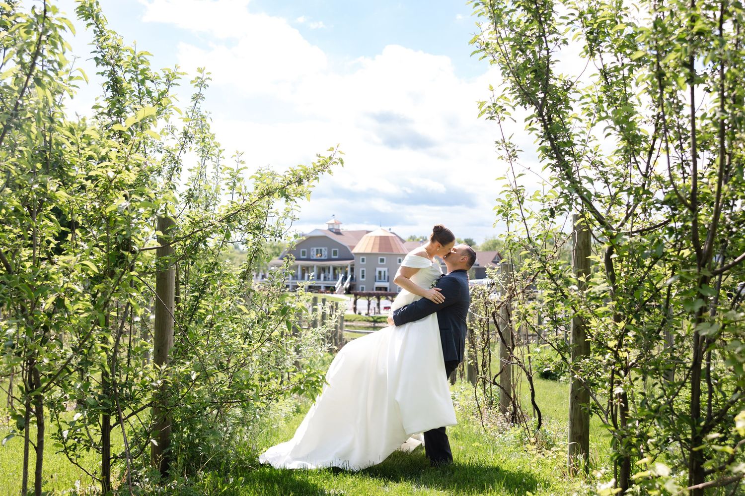 Bride and groom sharing a romantic kiss among lush green apple trees at an outdoor vineyard wedding venue.